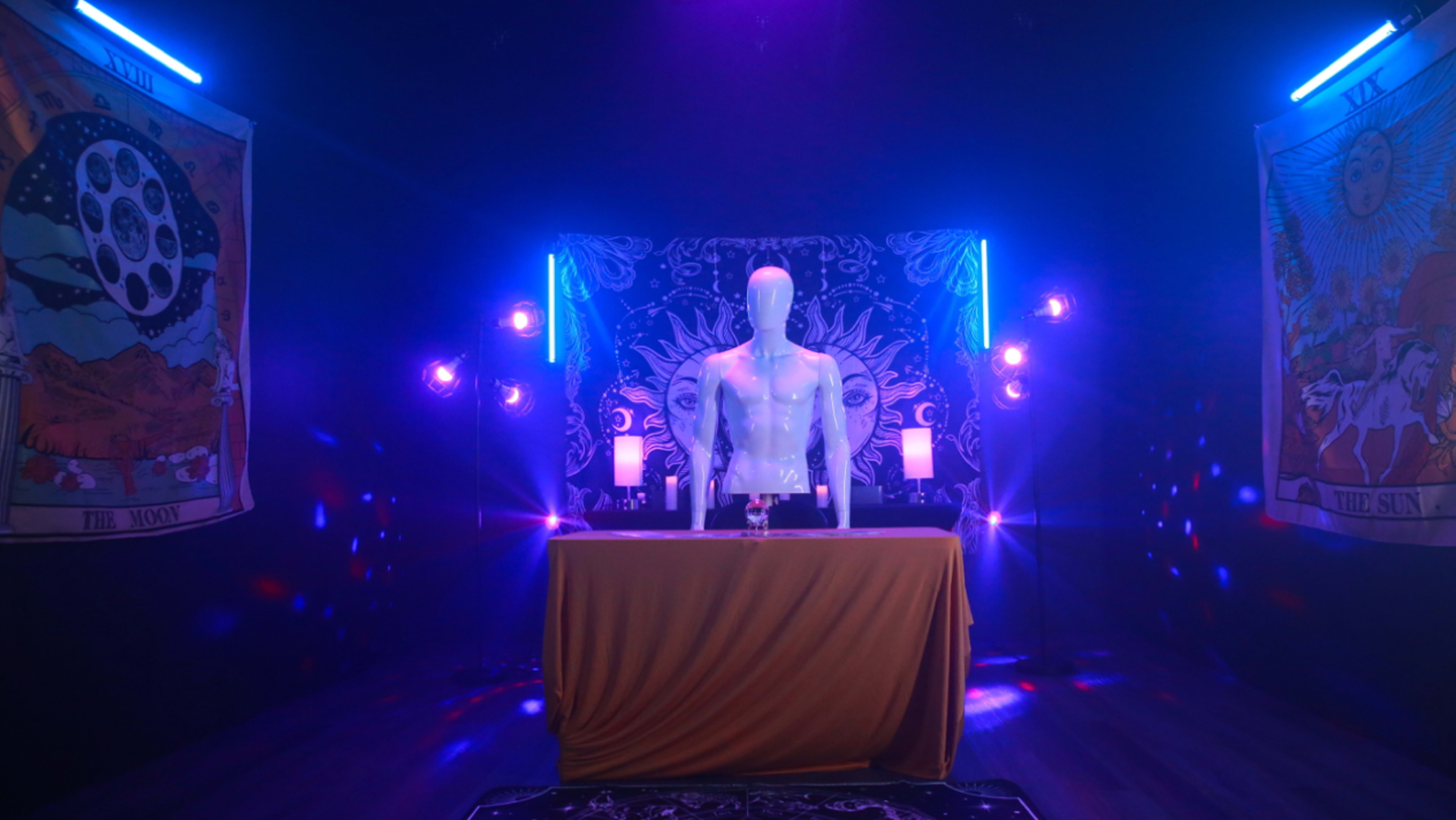 A mannequin stands on a table in a dimly lit room decorated with vibrant tapestries and colorful lighting.