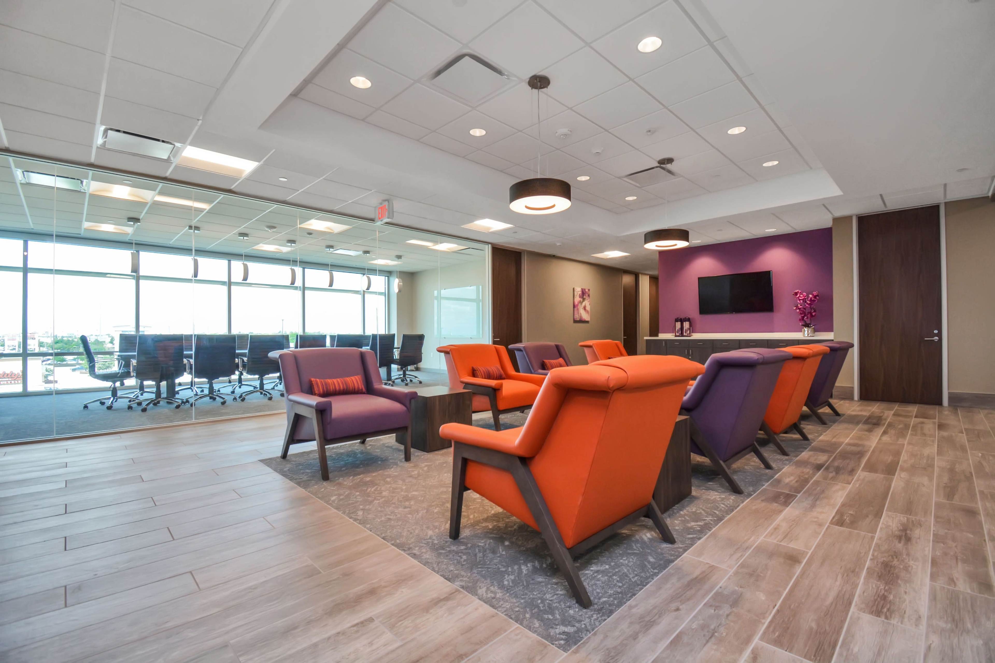 A modern office lounge area with orange and purple chairs, a bar-like counter, and a conference room visible through large windows.