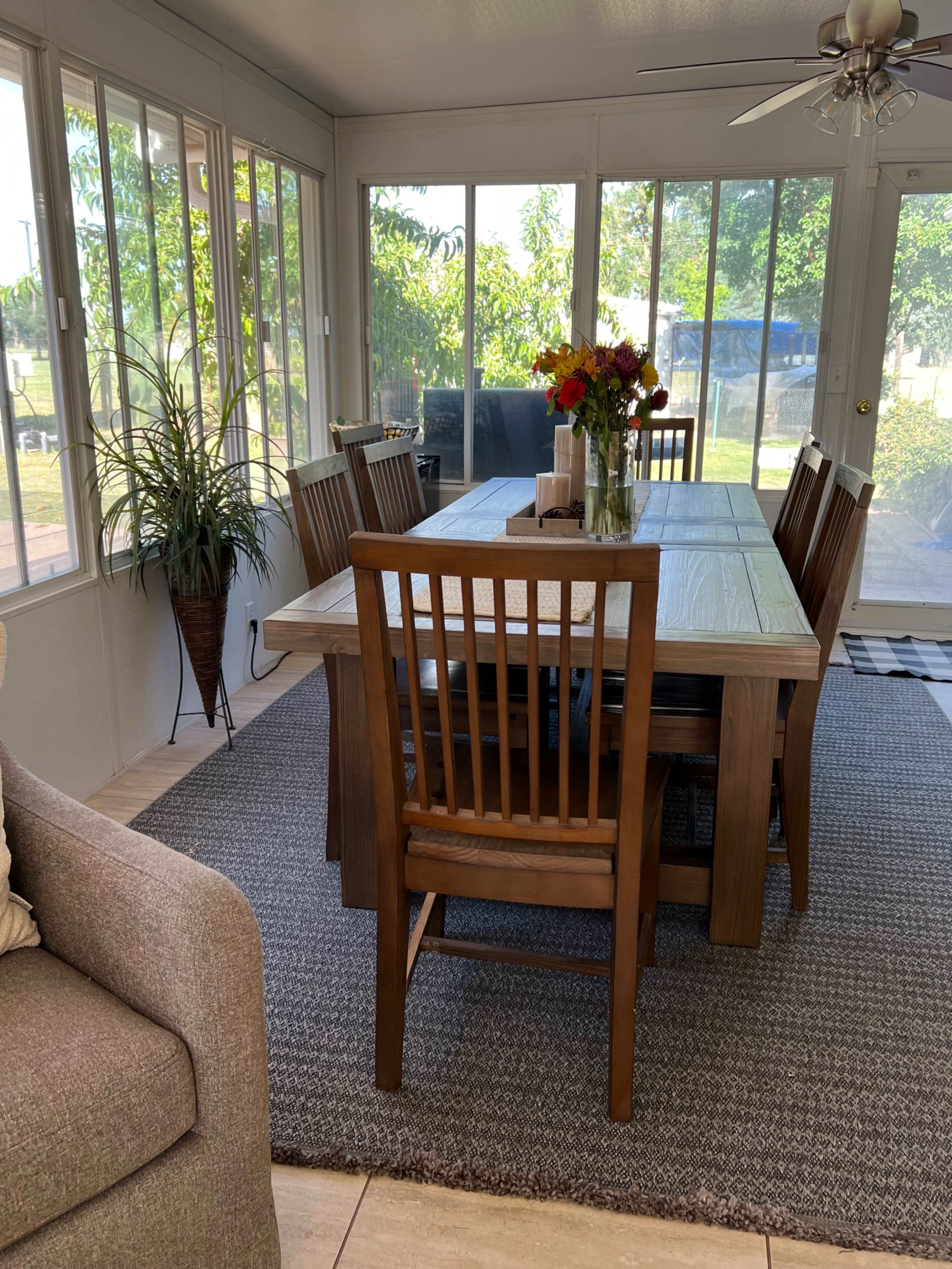 The image shows a well-lit sunroom with a wooden dining table surrounded by eight chairs, a vase of flowers on the table, and large windows allowing for natural light and a view of greenery outside.