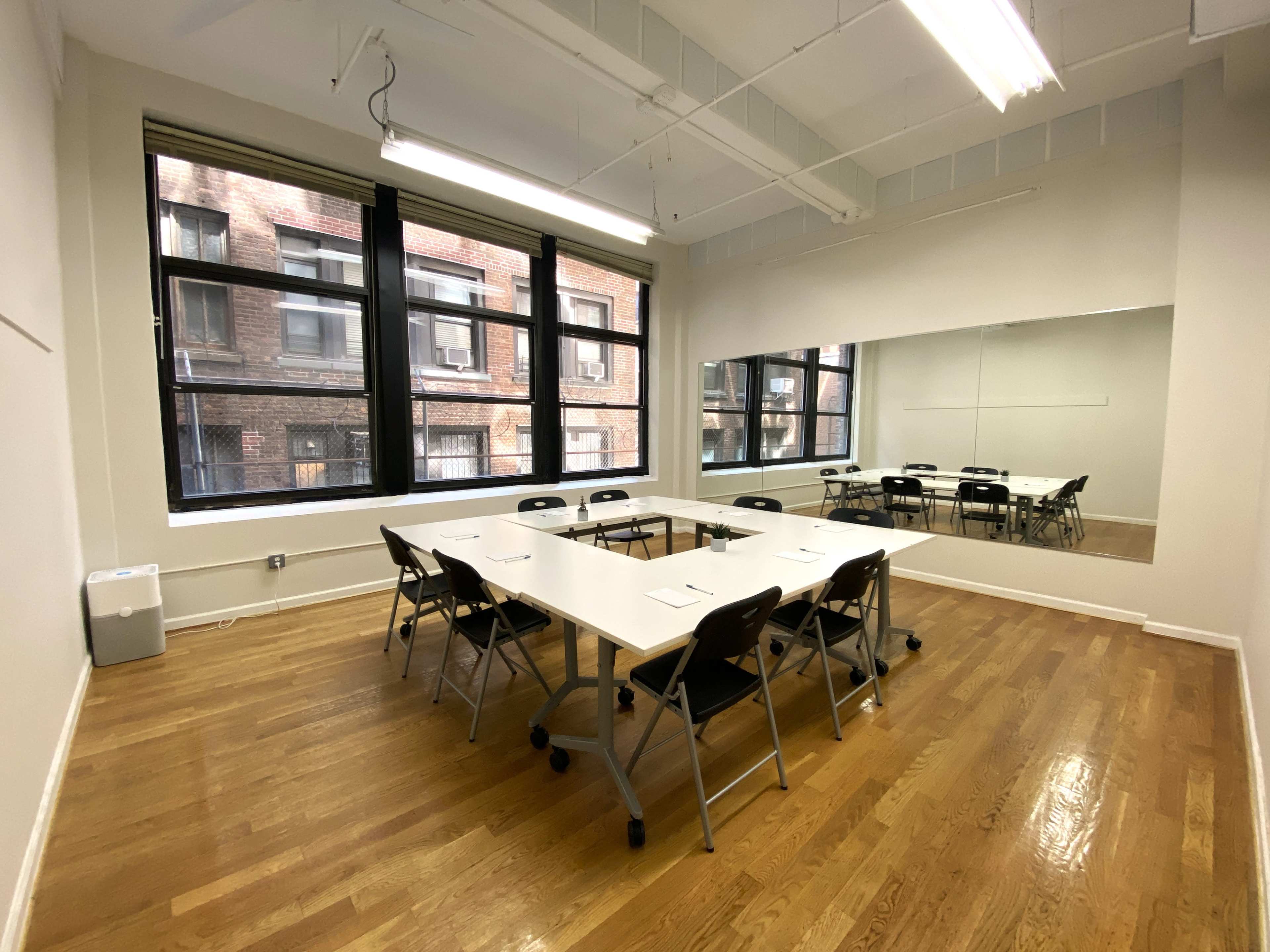 A bright, empty meeting room with a large table surrounded by chairs and large windows overlooking a brick building.