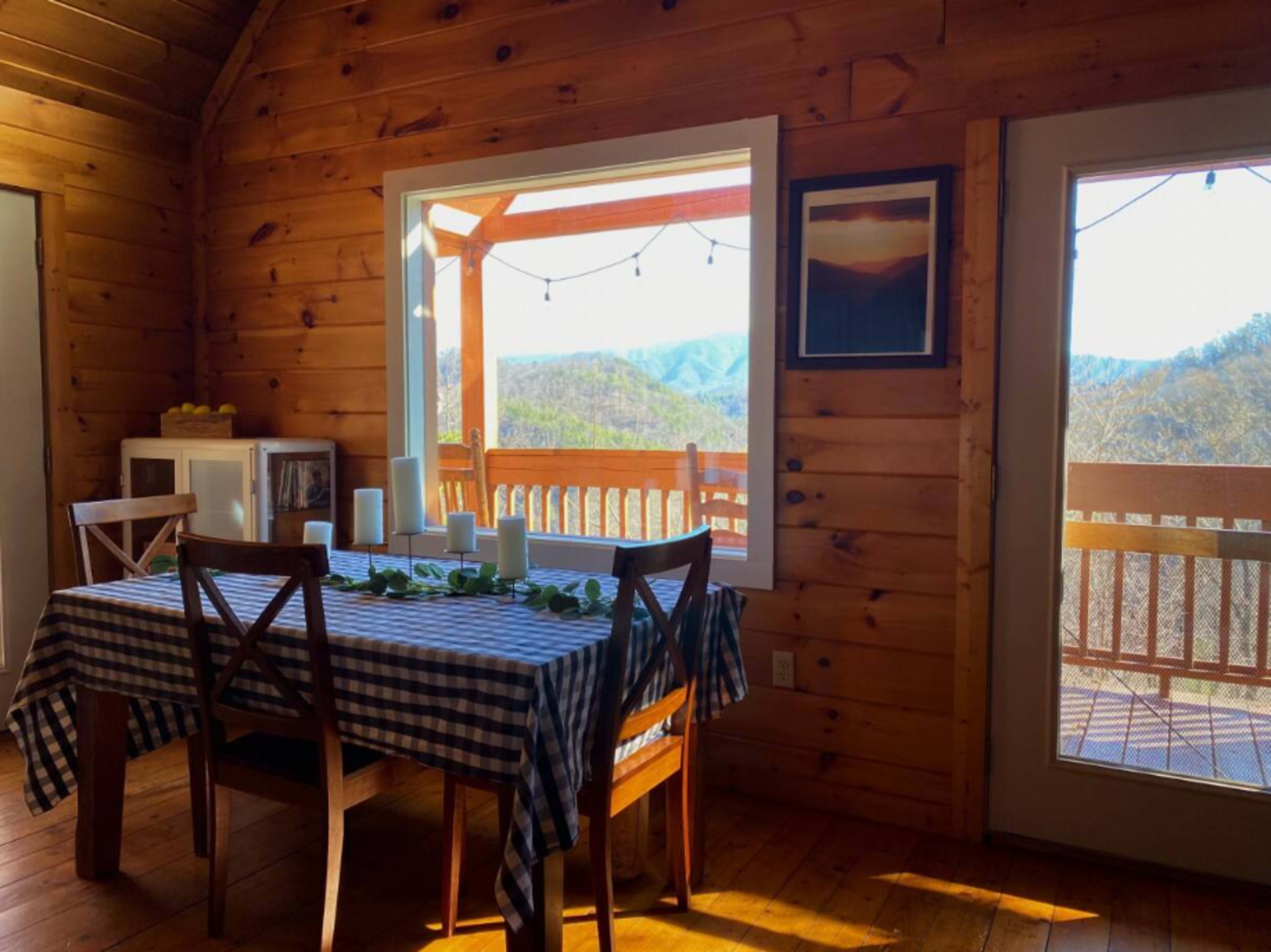 A dining table with a checkered tablecloth is set near a window that opens to a mountainous view.