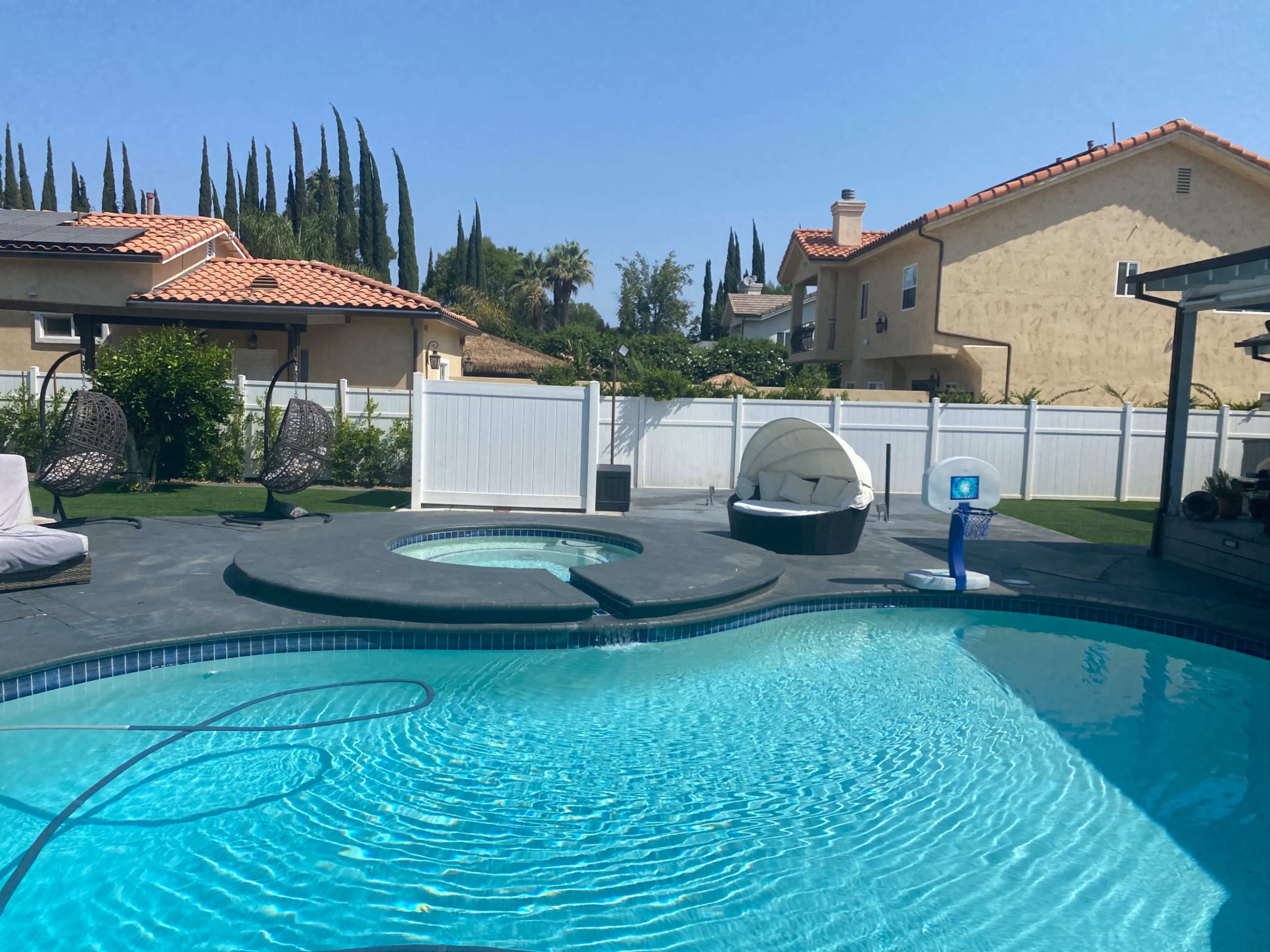 A clear blue swimming pool with a spa, surrounded by a neatly manicured yard and several tall trees in the background.