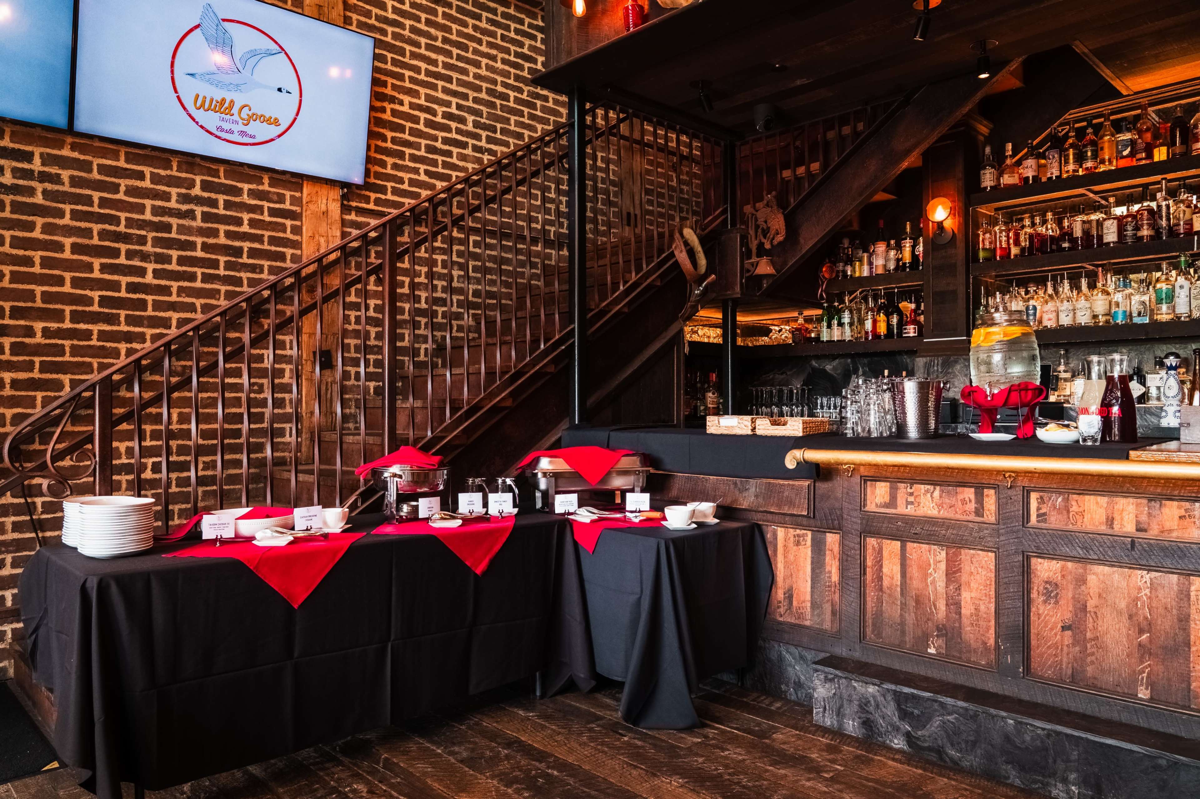 The image shows a restaurant bar area with a wooden counter, a variety of bottles on shelves, and a buffet table covered with black and red tablecloths.