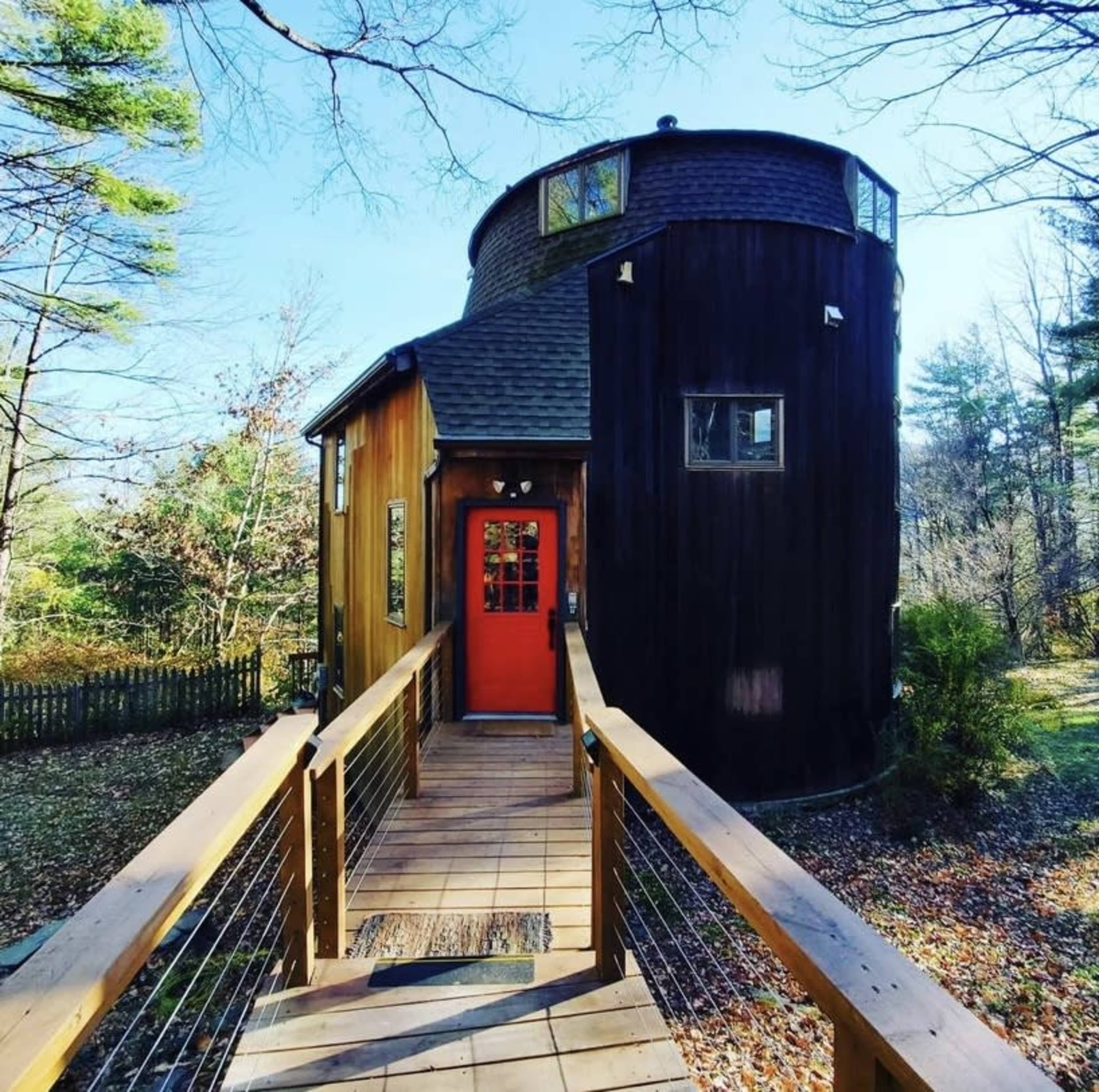 A round, two-story house with a red door is connected to a wooden deck surrounded by trees.