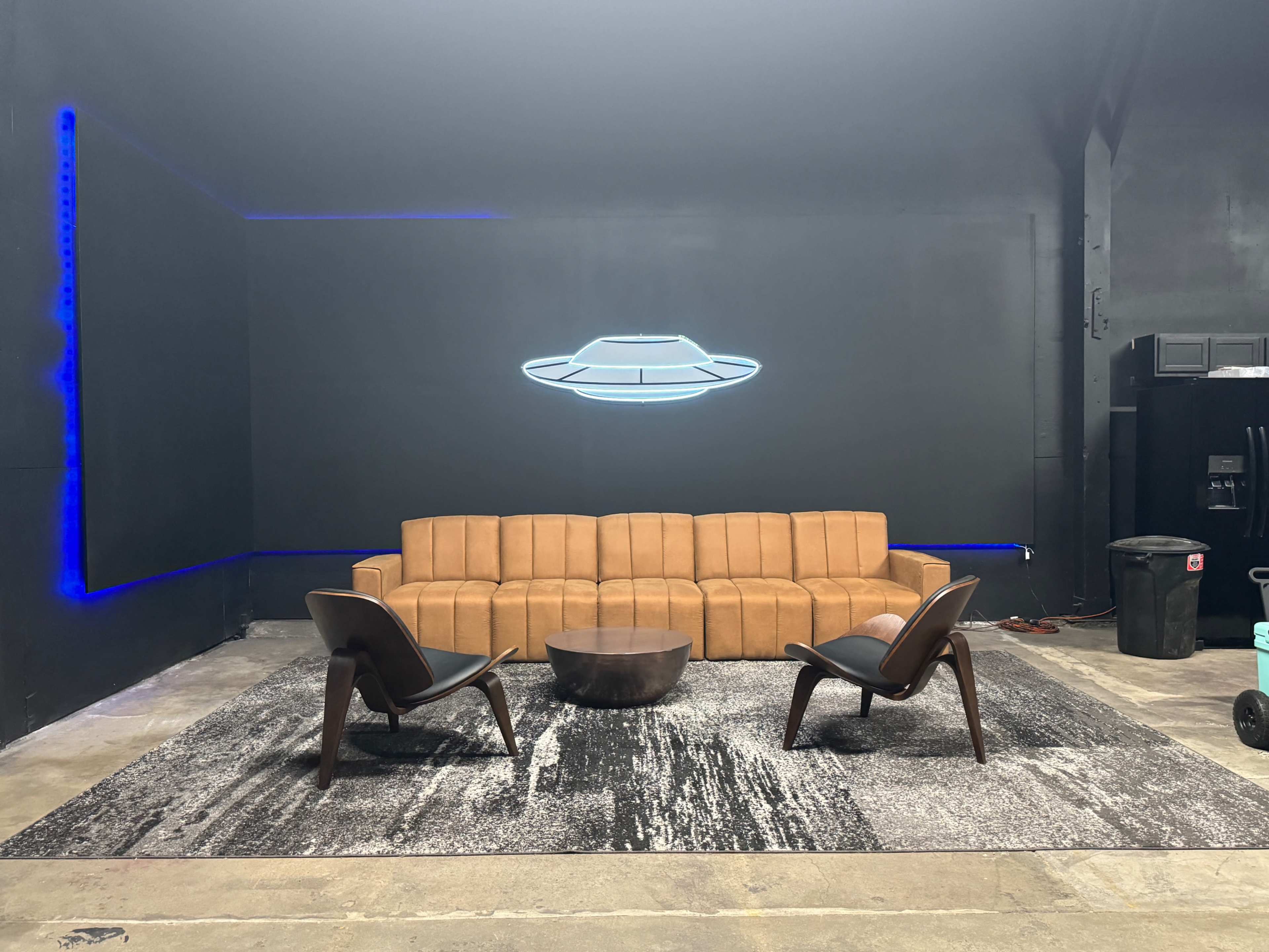 The image shows a modern interior featuring a tan sectional sofa, two black chairs, and a circular coffee table on a patterned rug, with a minimalist black wall illuminated by blue lighting and a futuristic hovering object design.