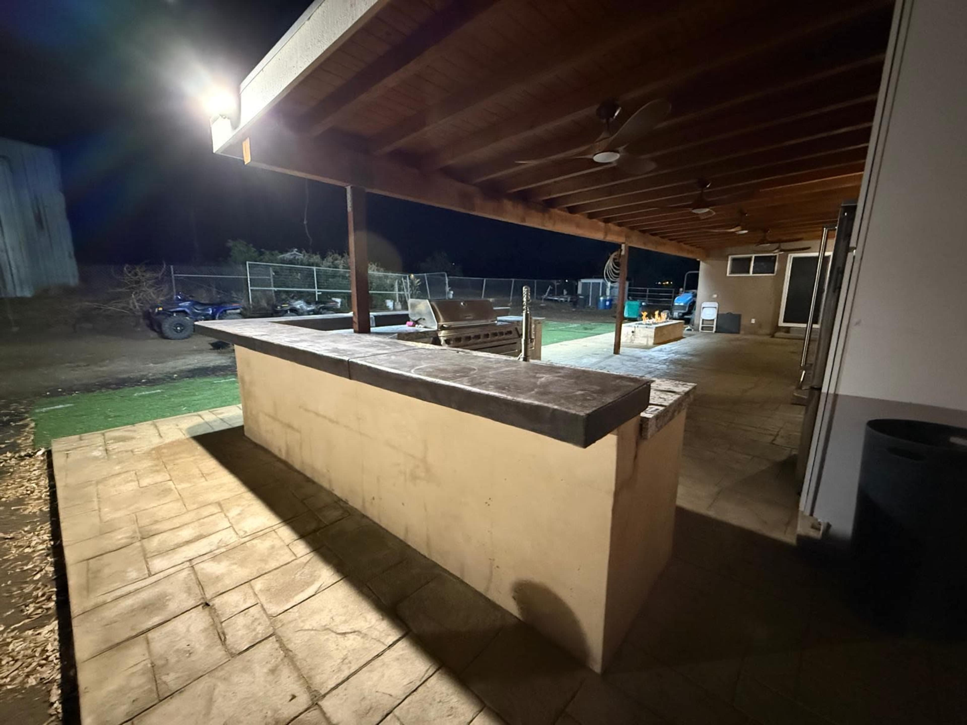 The image shows a dimly lit outdoor patio area featuring a large concrete bar with a wooden roof overhead.