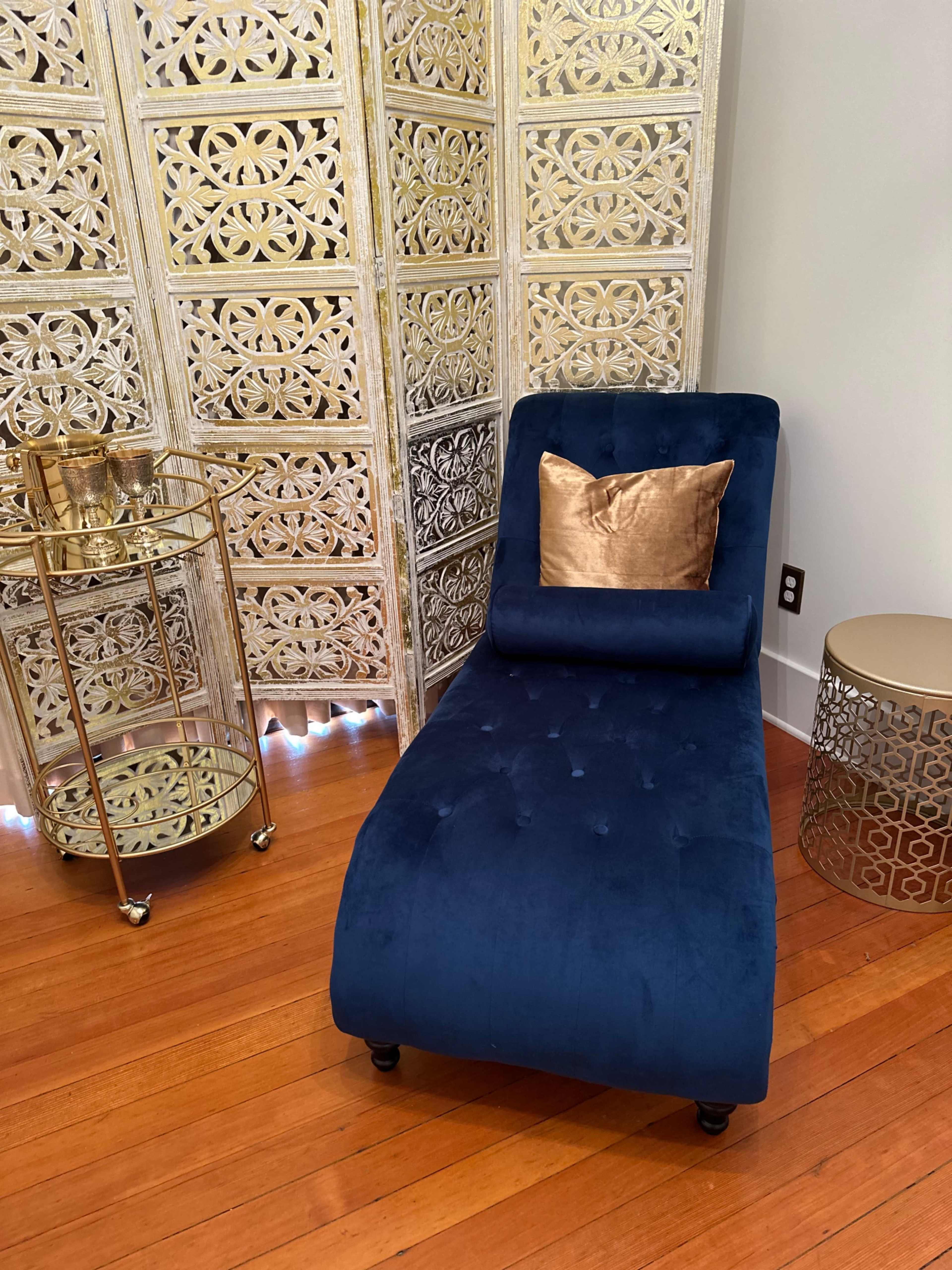 A blue chaise lounge with a gold pillow next to a decorative screen and a round gold side table.