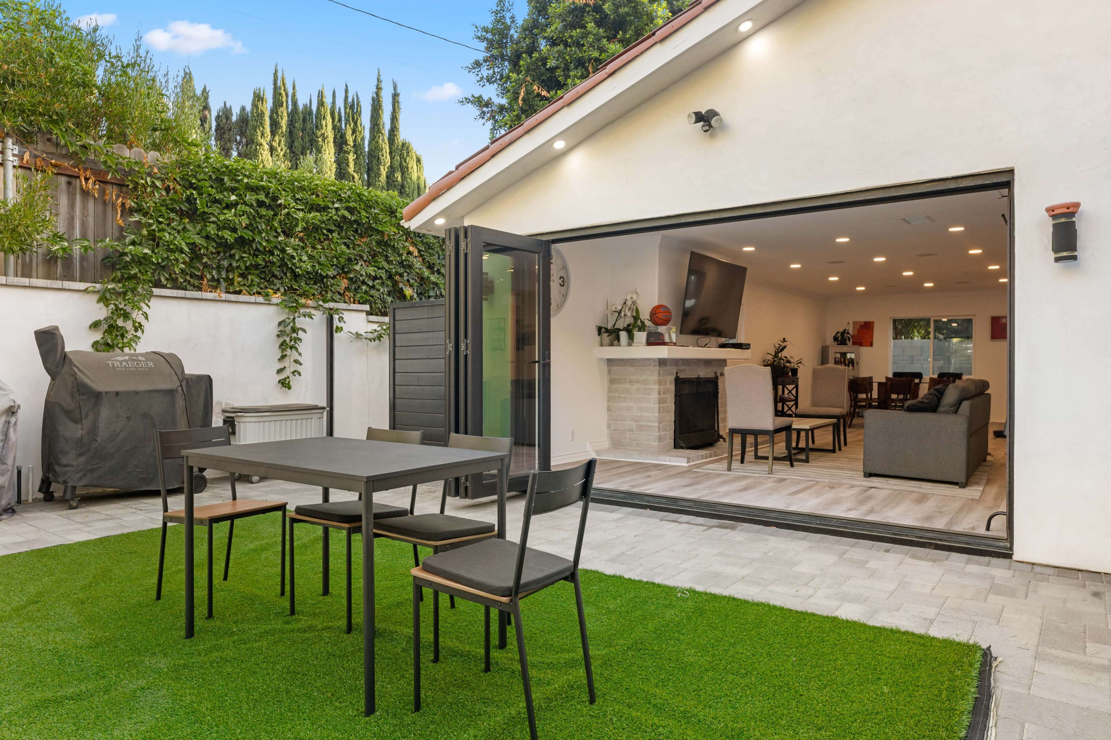 A modern outdoor patio features a black dining table with four chairs, a lush green lawn, and a spacious open living area visible through a large sliding door.