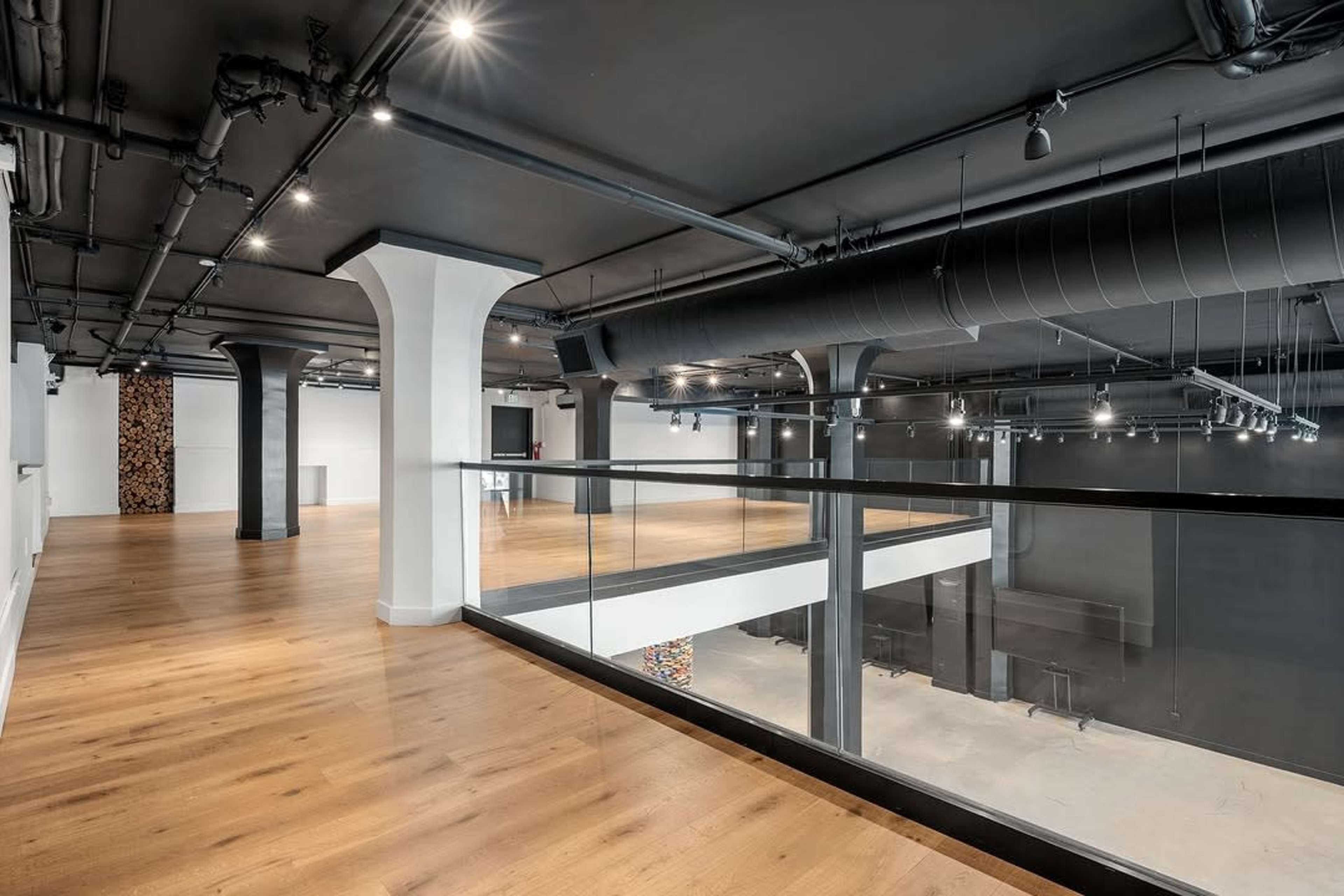 The image shows a modern, spacious interior of a commercial space with wooden flooring, high ceilings, and exposed ductwork, featuring a glass railing on a balcony overlooking an open area below.