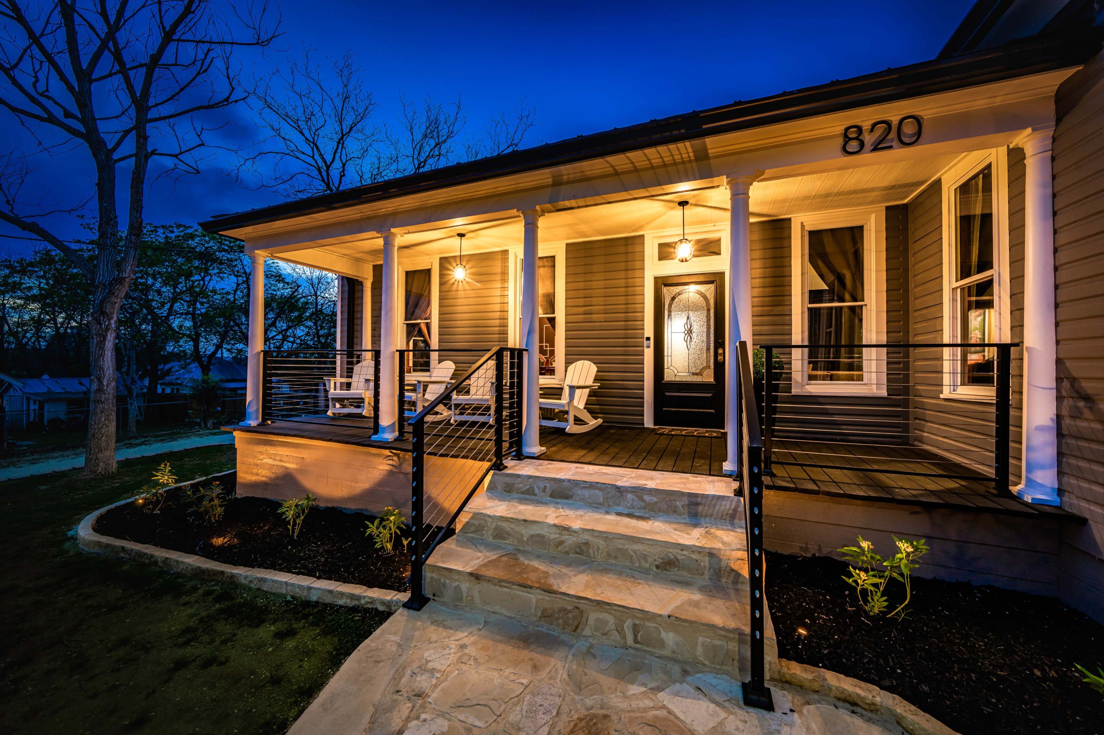 A well-lit porch with white rocking chairs and a stone walkway leading up to a house at dusk.