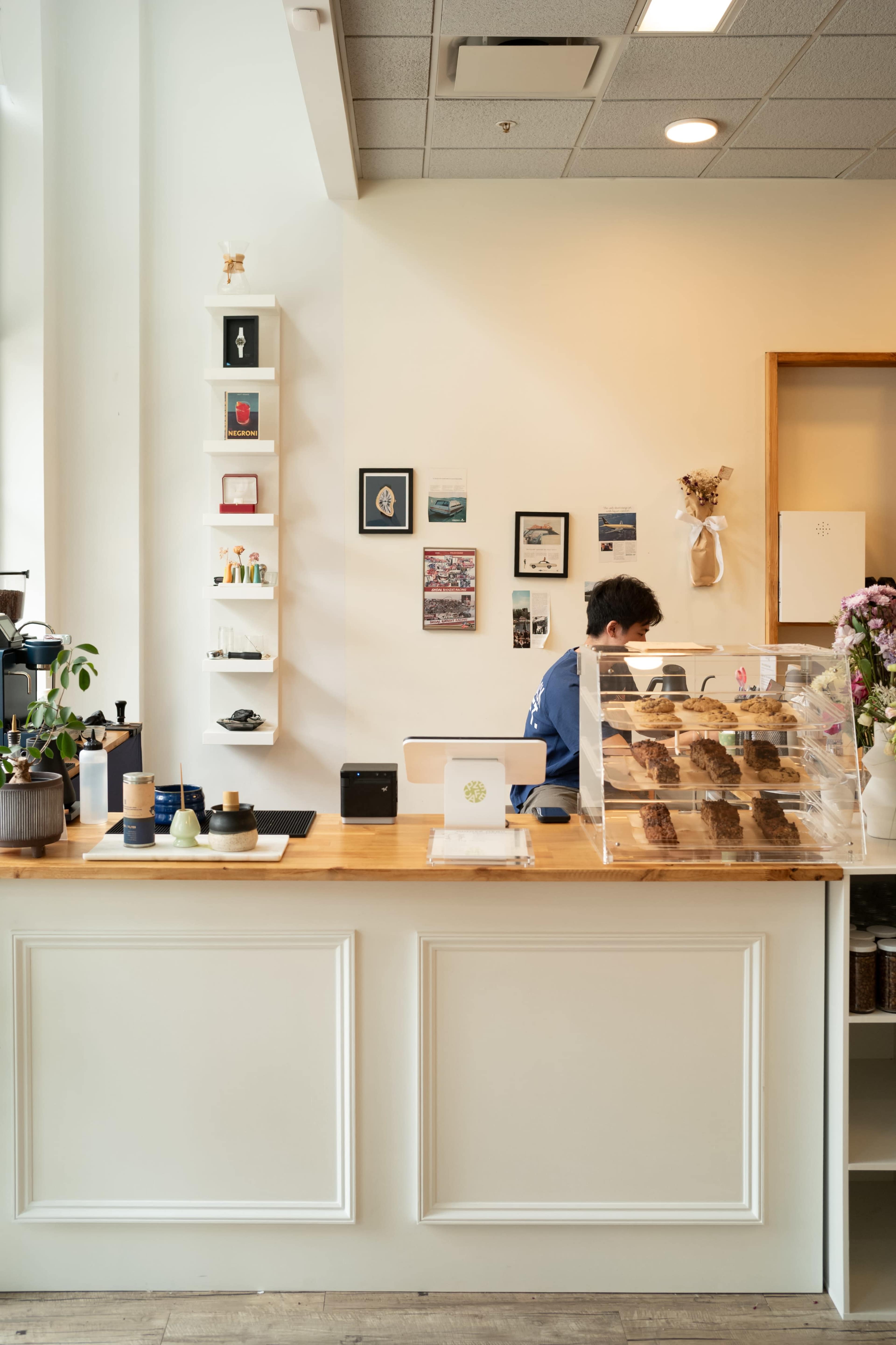 A barista works behind the counter of a cozy café, surrounded by decorative shelves and framed photos on the wall.