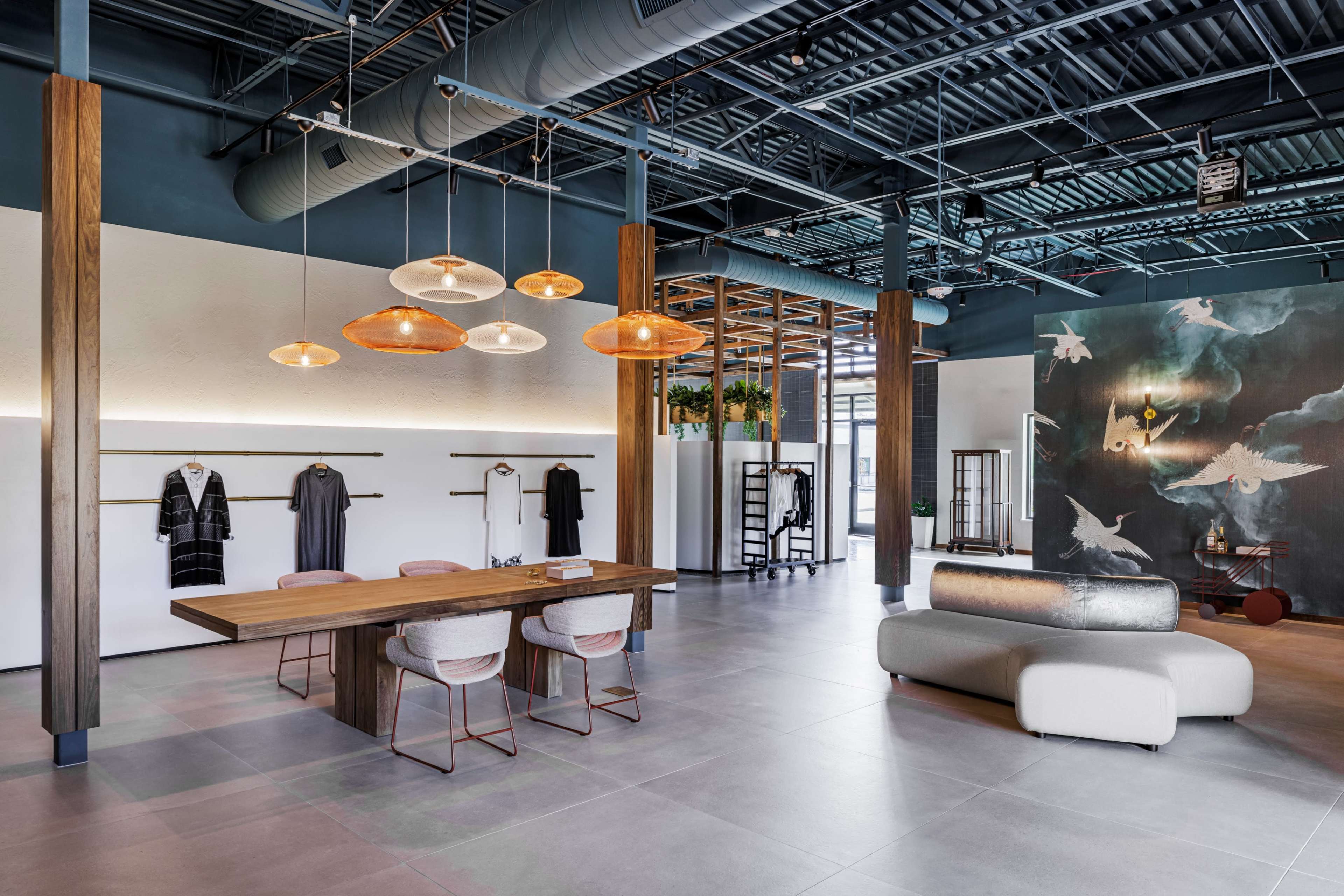 The image shows a modern office space featuring a large wooden table with chairs, stylish lighting fixtures, and clothing displays along the walls.