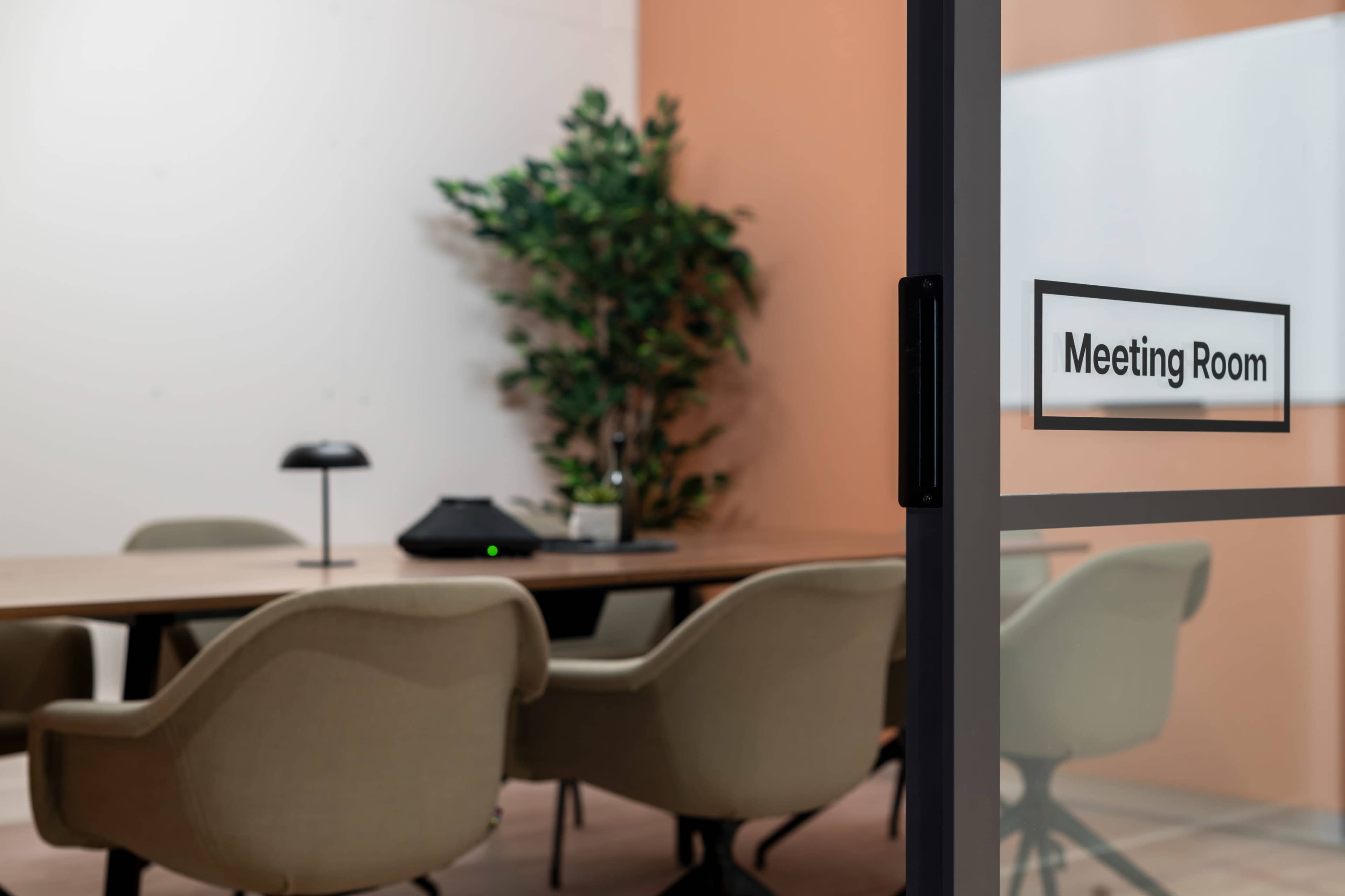 A glass door with a "Meeting Room" sign leads to a conference room featuring a wooden table surrounded by beige chairs and a potted plant in the background.