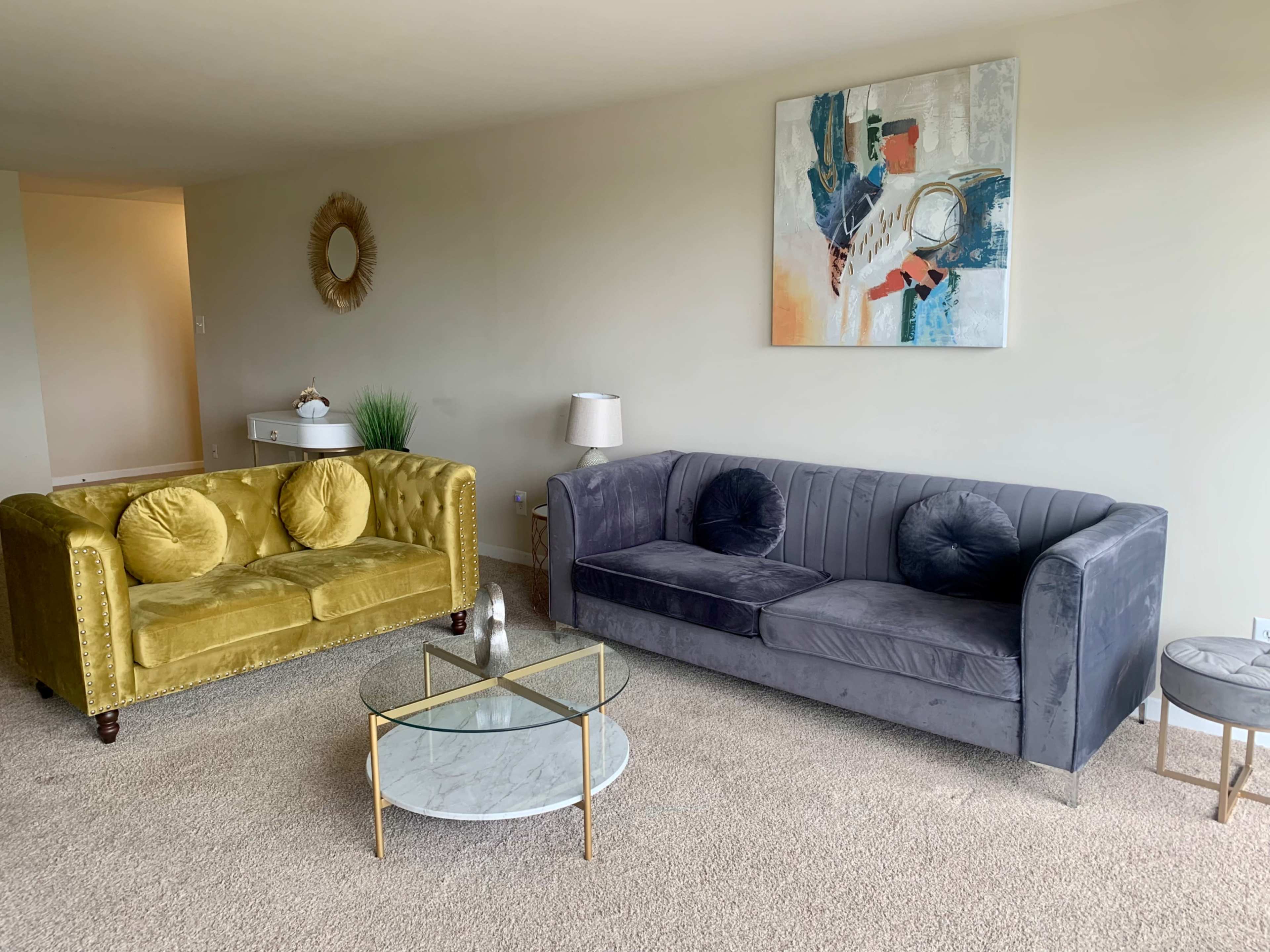 A living room with a yellow velvet couch, a gray upholstered couch, a glass coffee table, and a decorative wall art piece.
