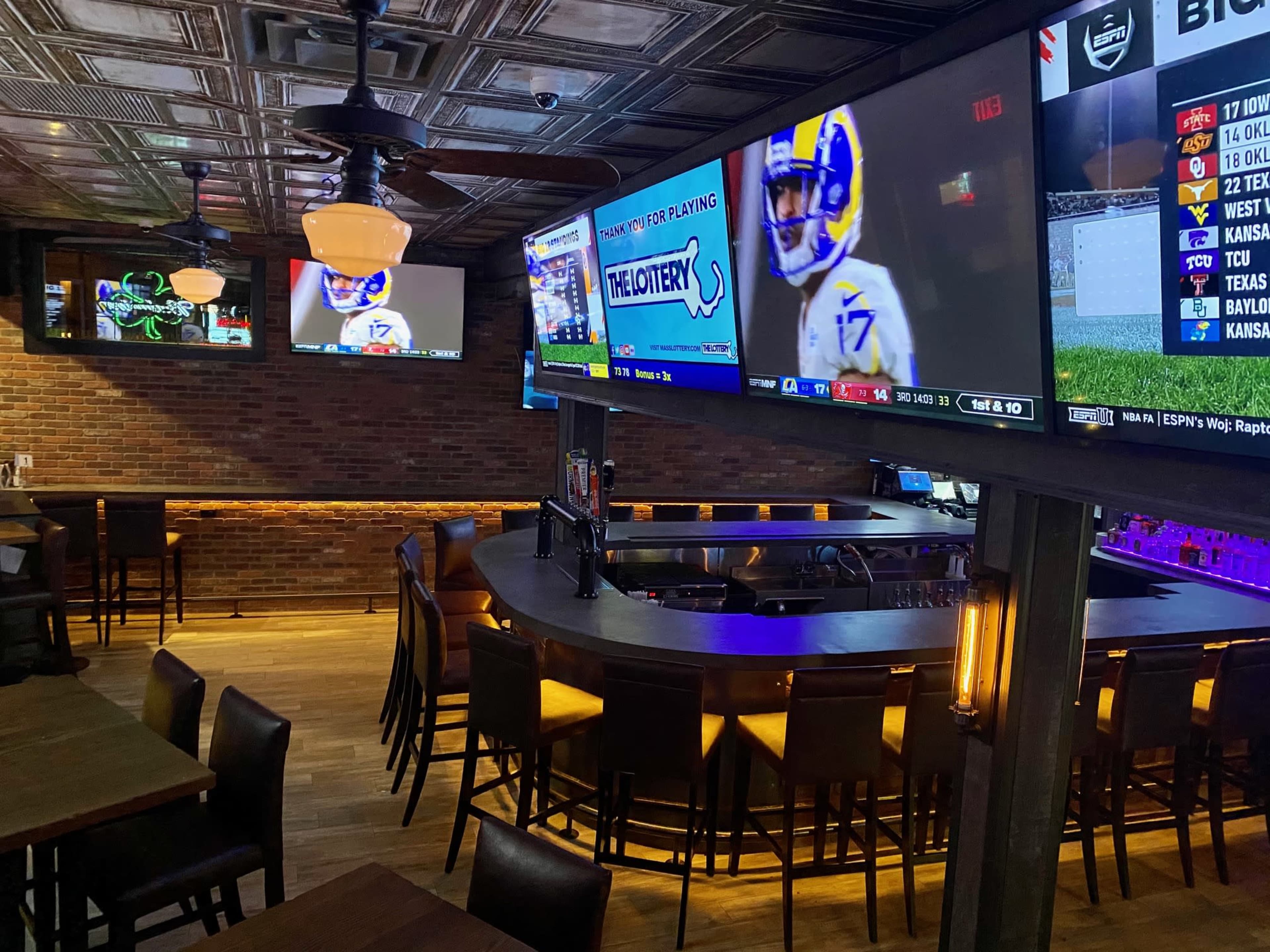 A sports bar interior with multiple televisions displaying a football game, wooden tables, and a curved bar area under industrial-style lighting.