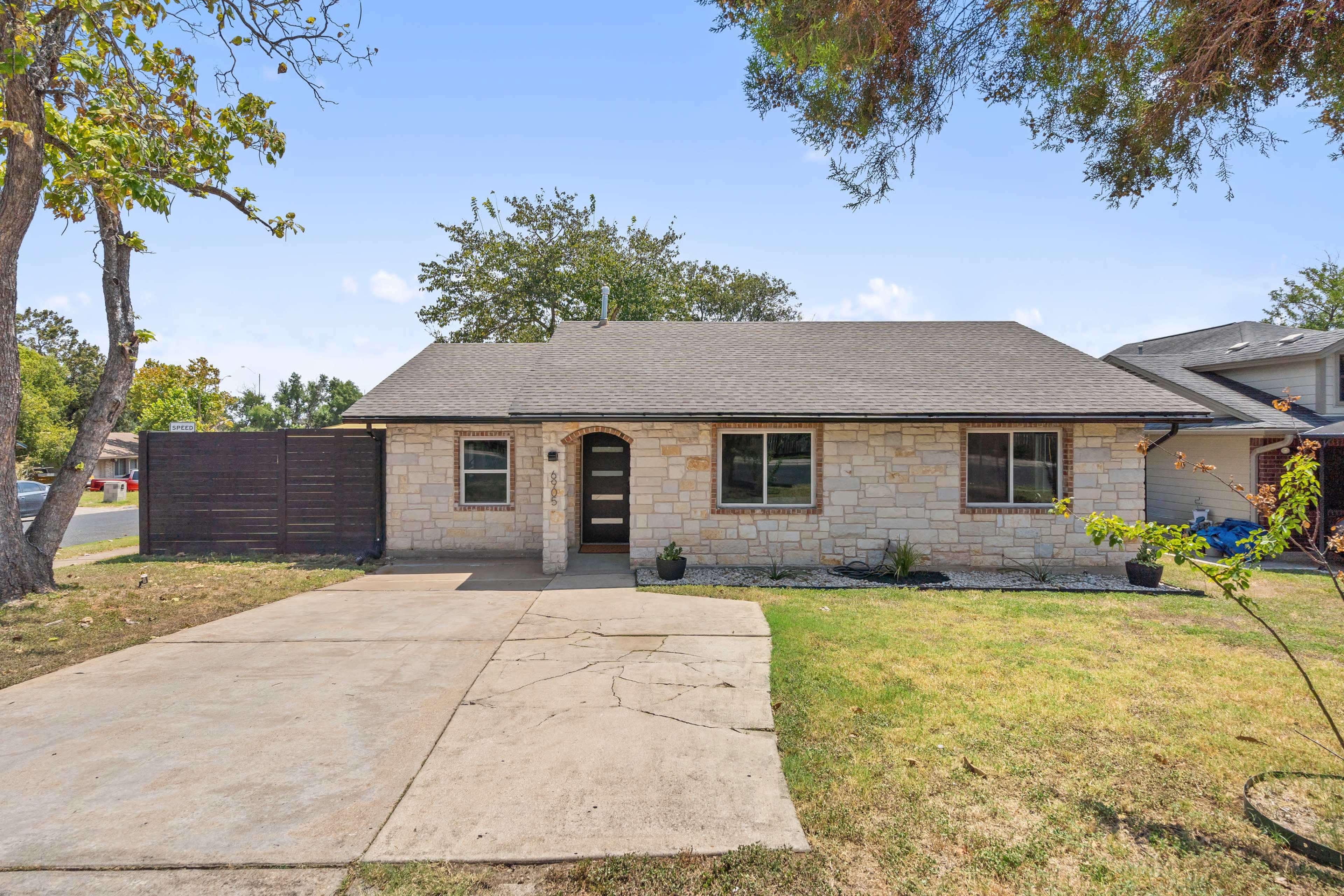 The image shows a single-story stone house with a sloped roof and a small front yard, situated near a street.