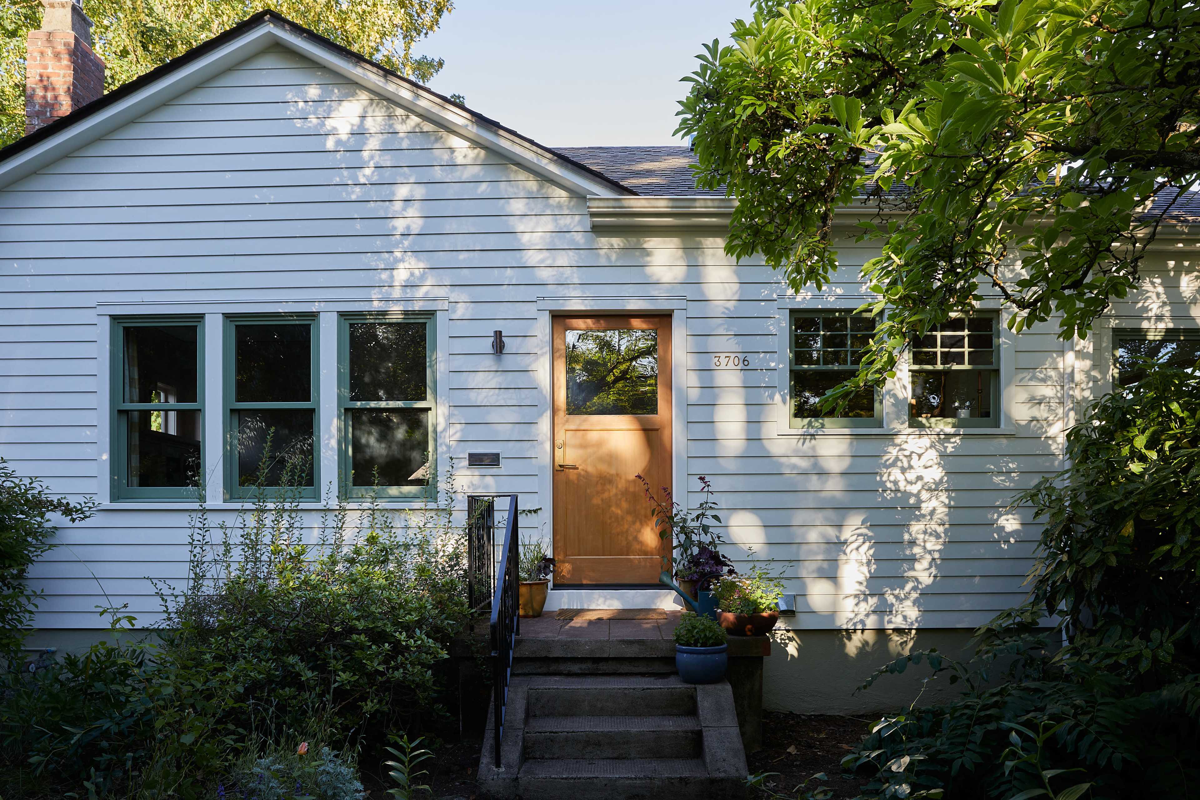 A light-colored house with a wooden front door and several windows is framed by greenery and shadows.
