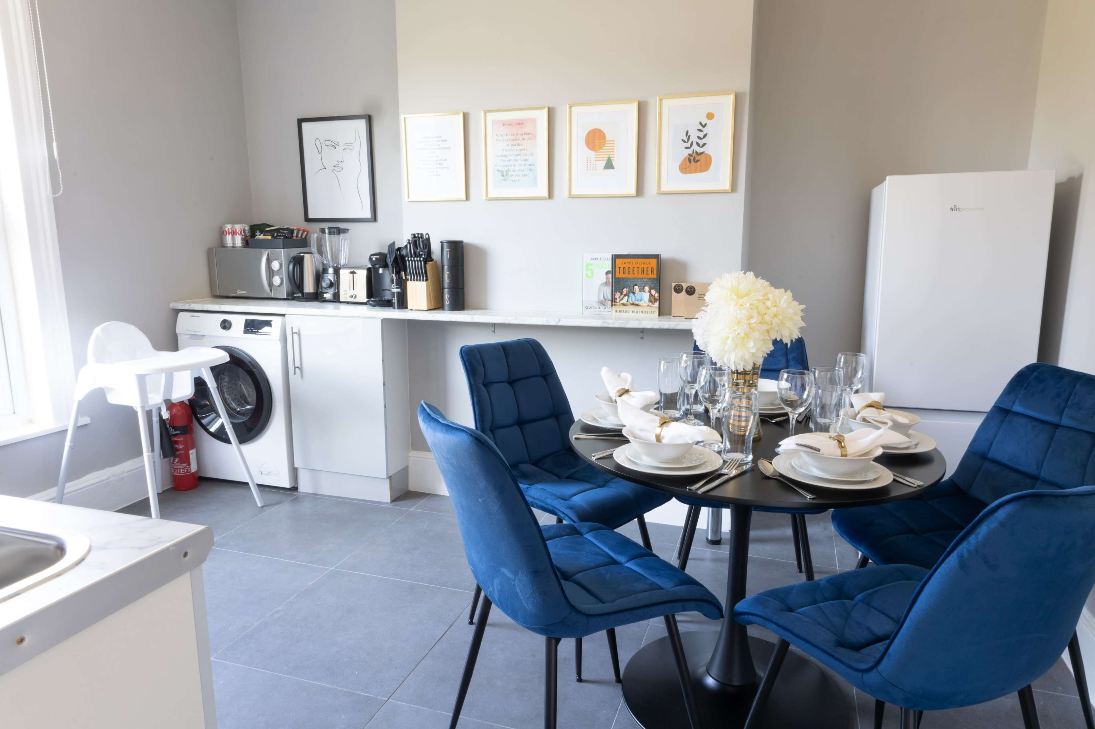 A modern kitchen with a round black dining table set for four and equipped with a washing machine, coffee maker, and a white refrigerator.