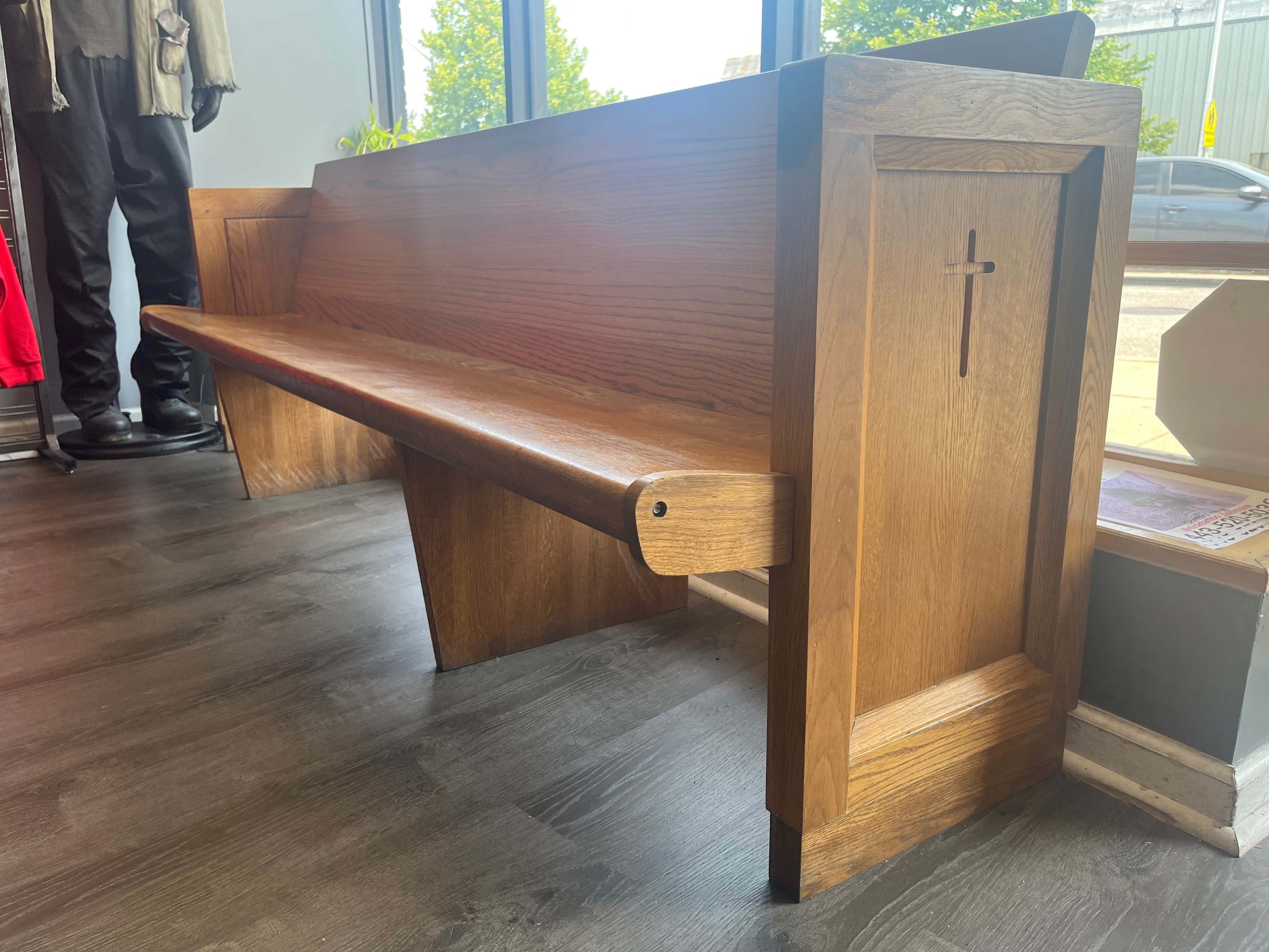 A wooden church pew with a cross symbol on its side, positioned next to a large window in a room with dark flooring.