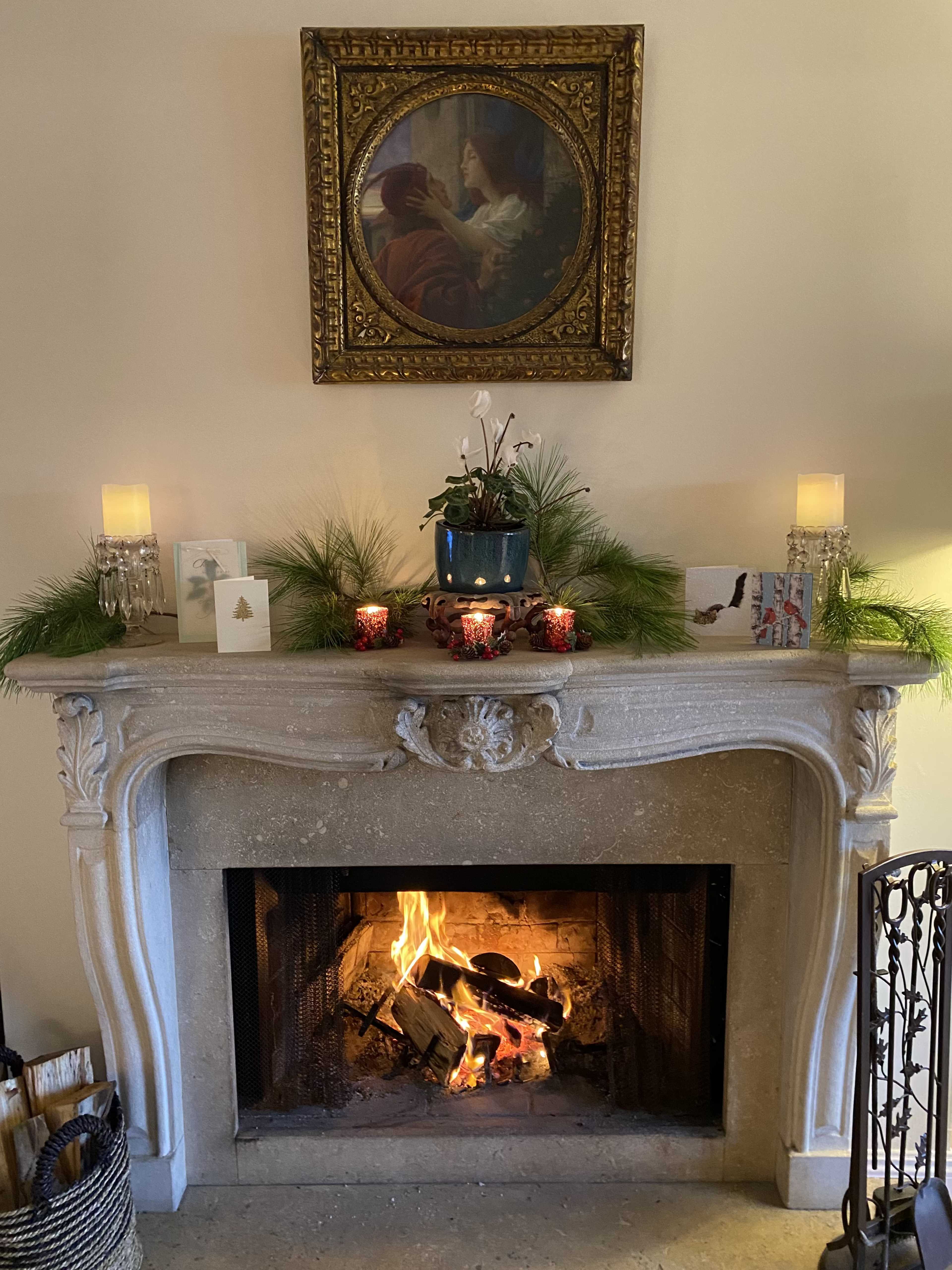 A stone fireplace with a lit fire is centered beneath a framed painting, surrounded by candles, greenery, and decorative items on the mantel.