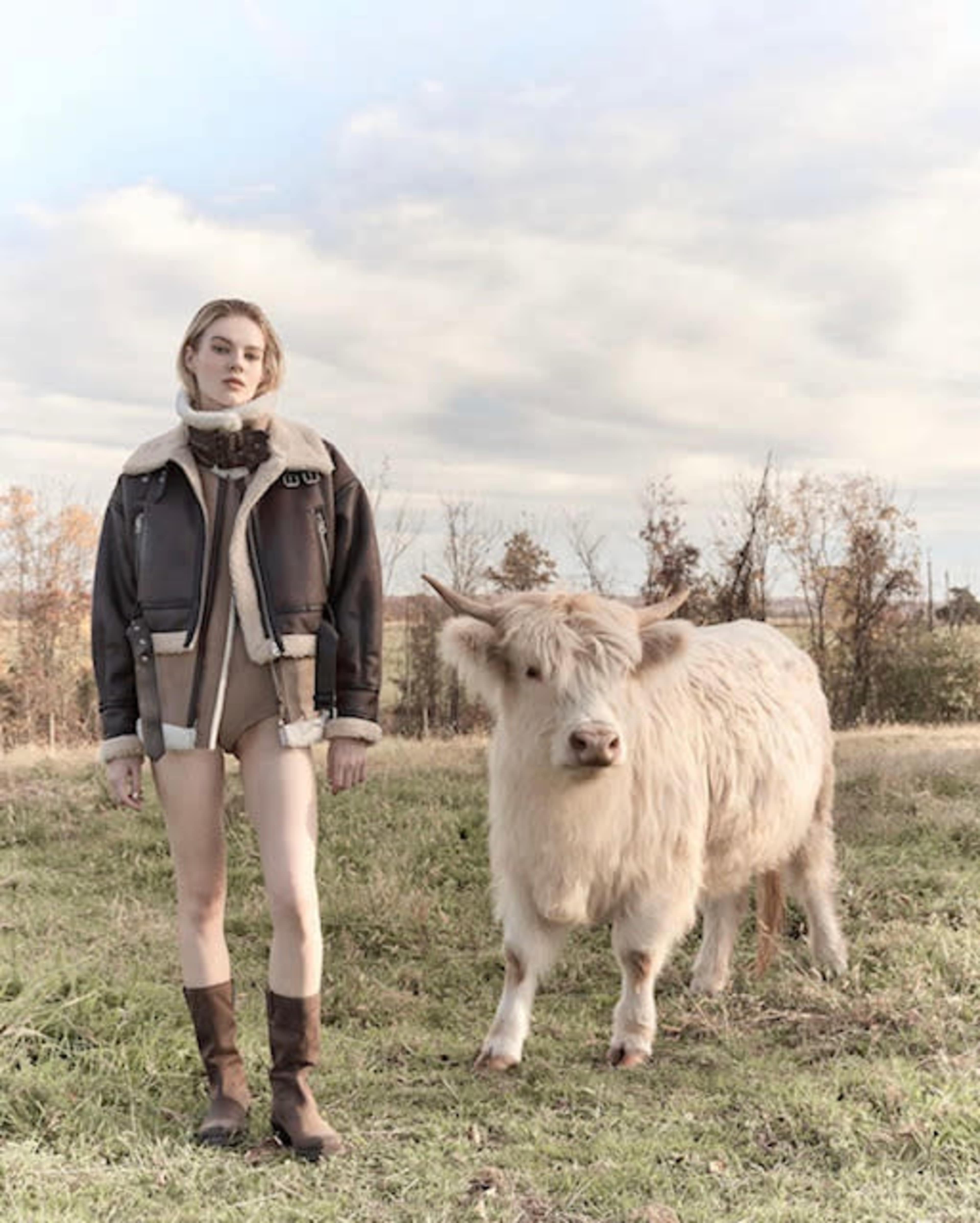 A young woman stands in a field next to a fluffy white cow under a cloudy sky.