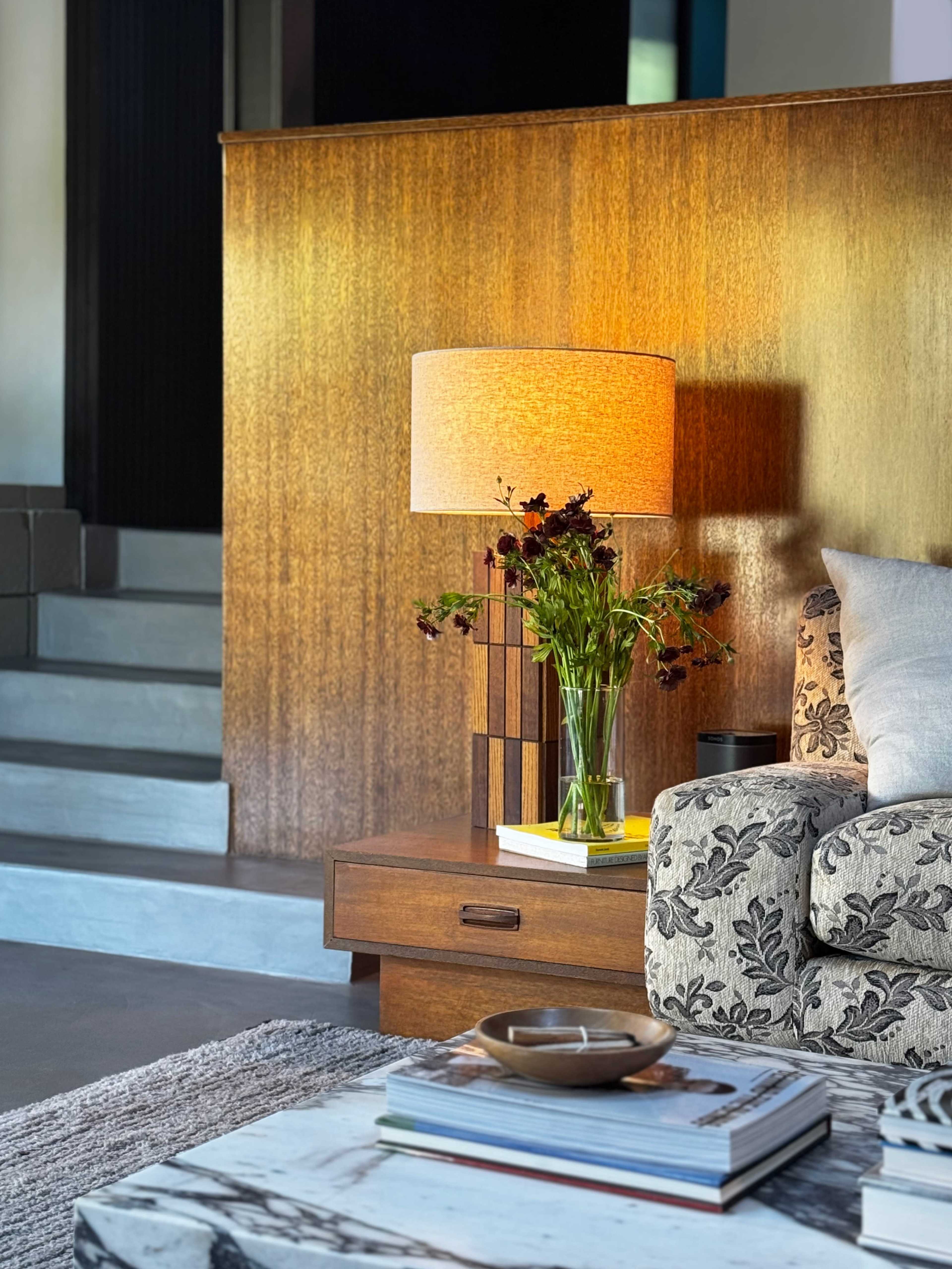 A living room scene features a patterned couch alongside a wooden side table with a lamp and a vase of flowers, with stairs visible in the background.