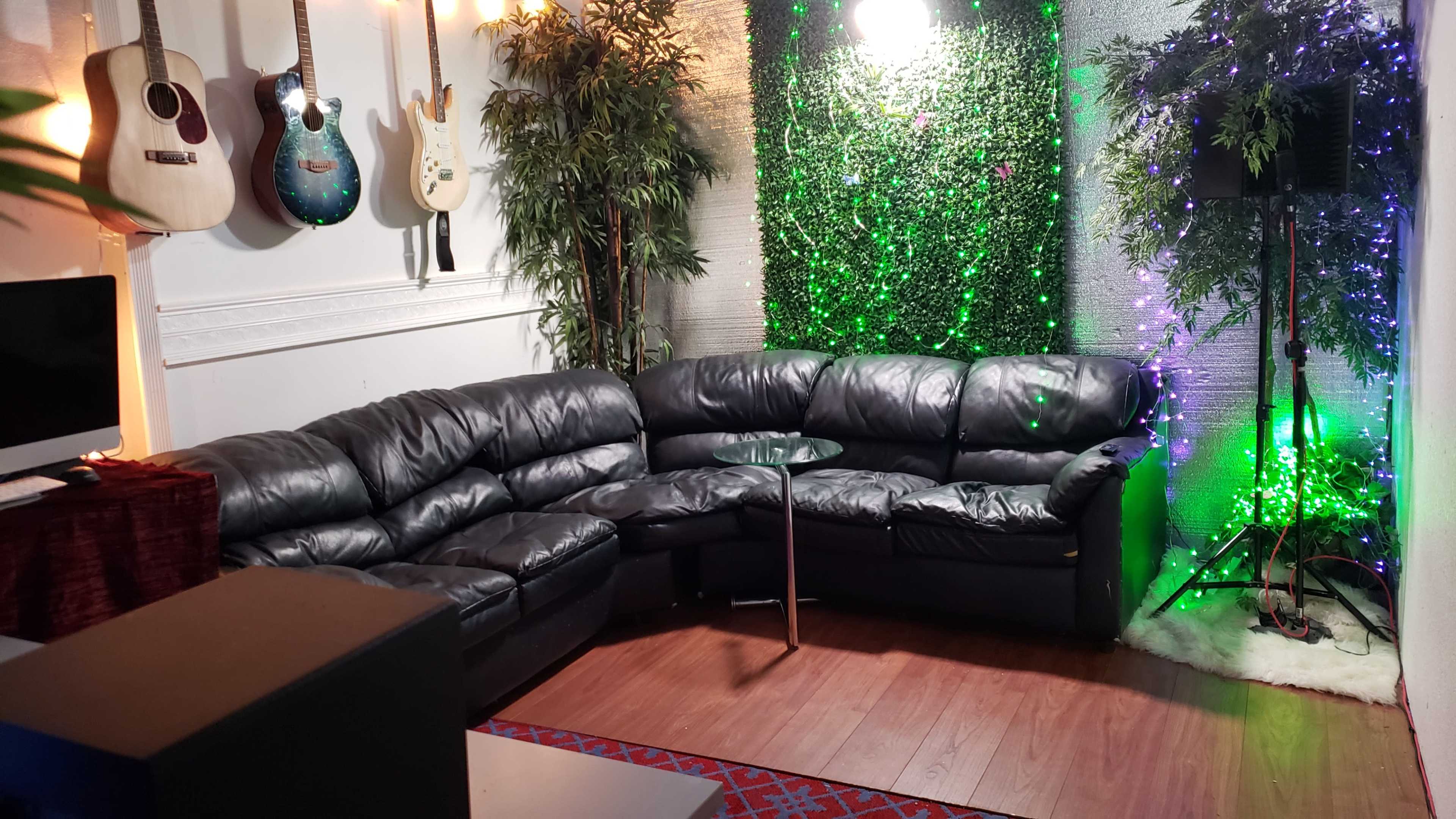 A cozy lounge area with a black leather sectional sofa, a glass table, illuminated plants, and guitars mounted on the wall.