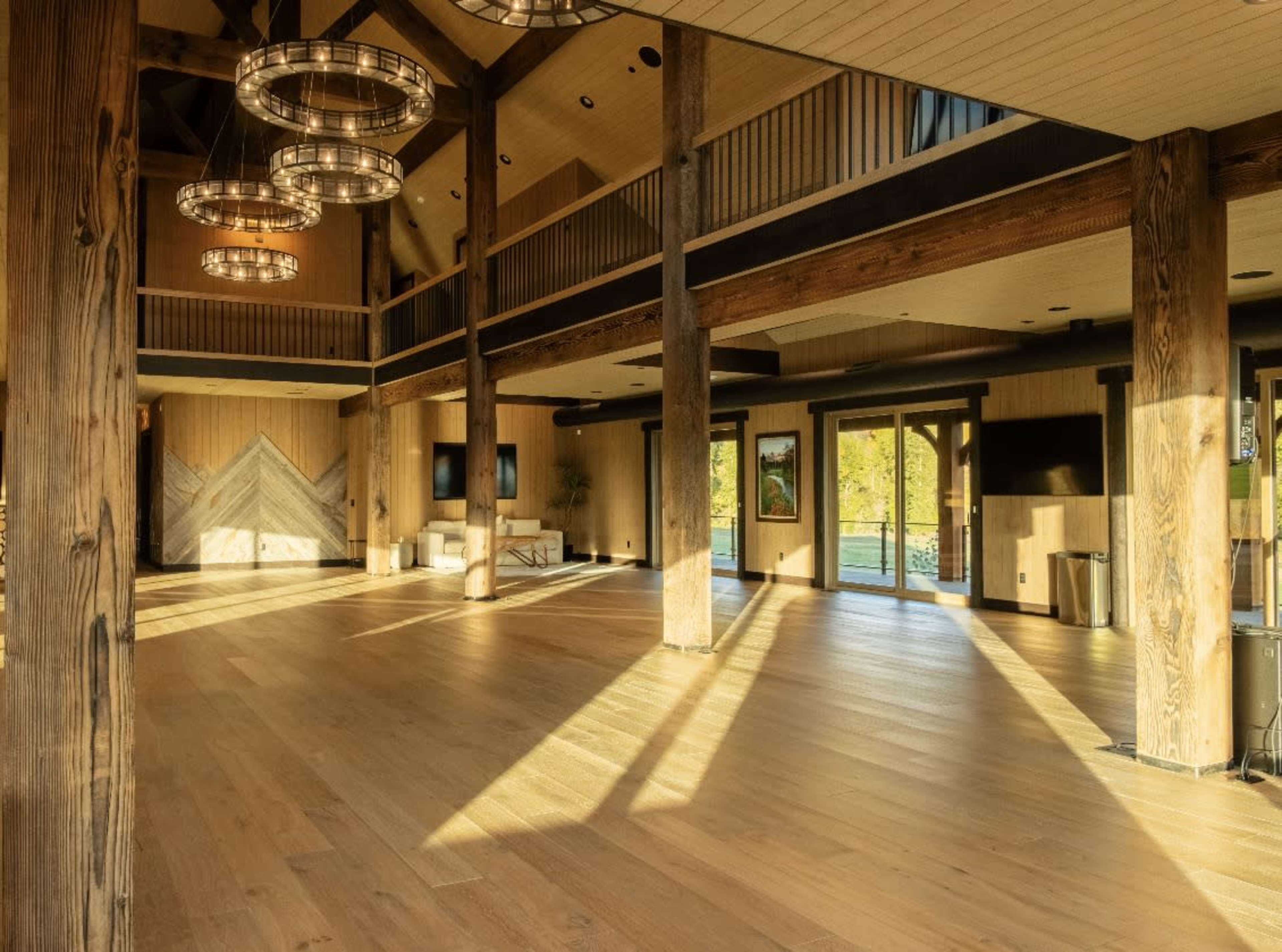The image shows a spacious interior of a building with wooden beams, large windows, and warm lighting casting shadows on the wooden floor.