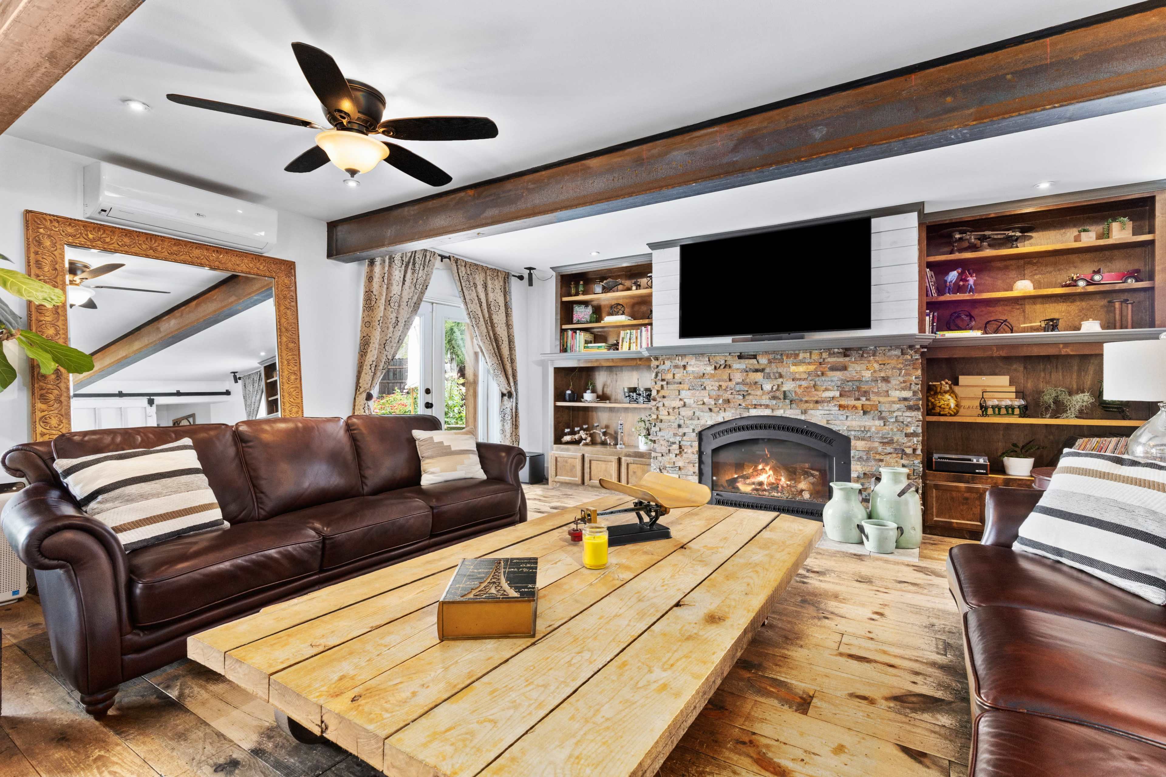 A modern living room with a stone fireplace, a wooden coffee table, and brown leather sofas, illuminated by natural light.