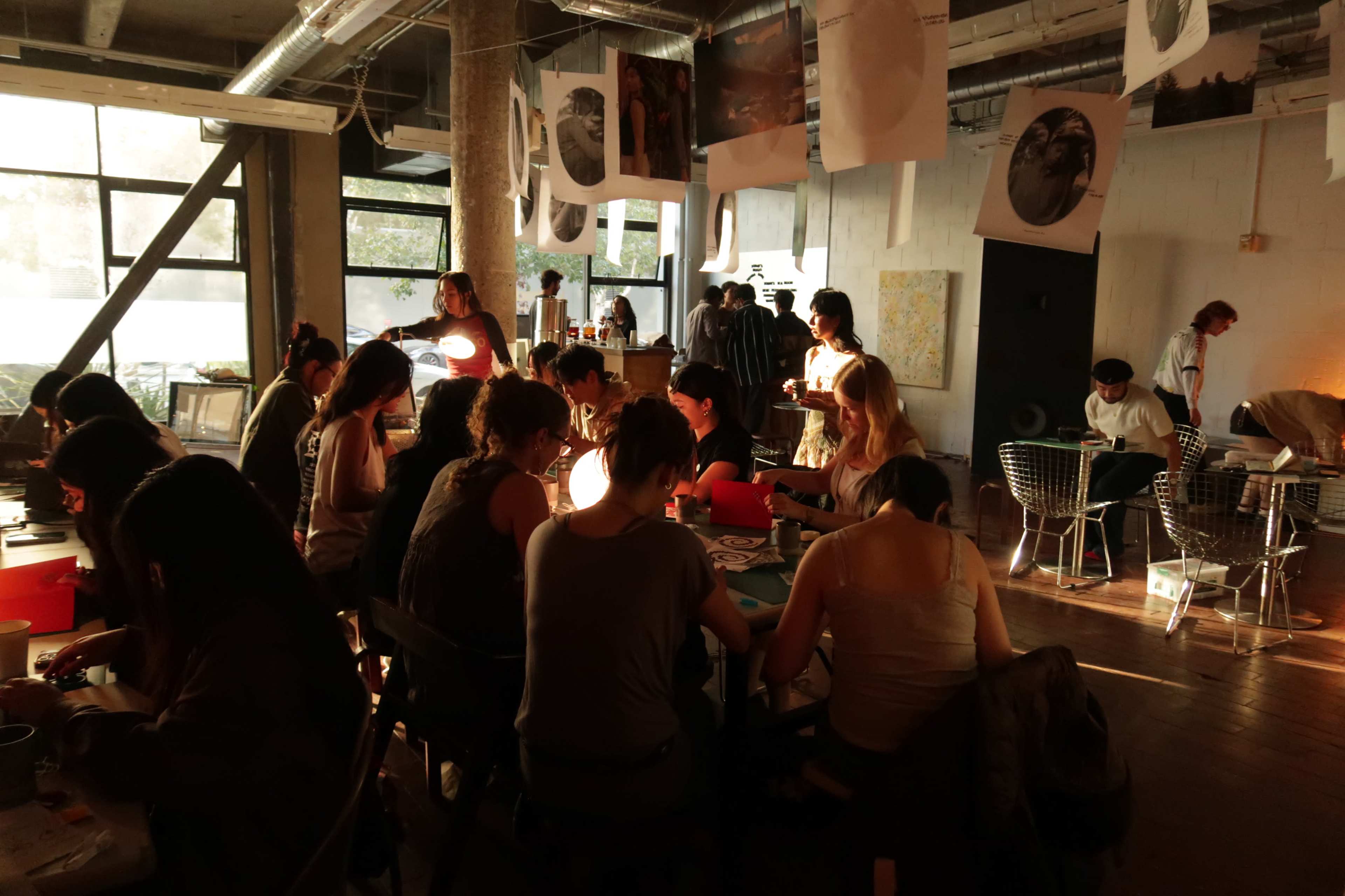 A group of people is engaged in activities around tables in a dimly lit, creative workspace, with several standing and interacting in the background.