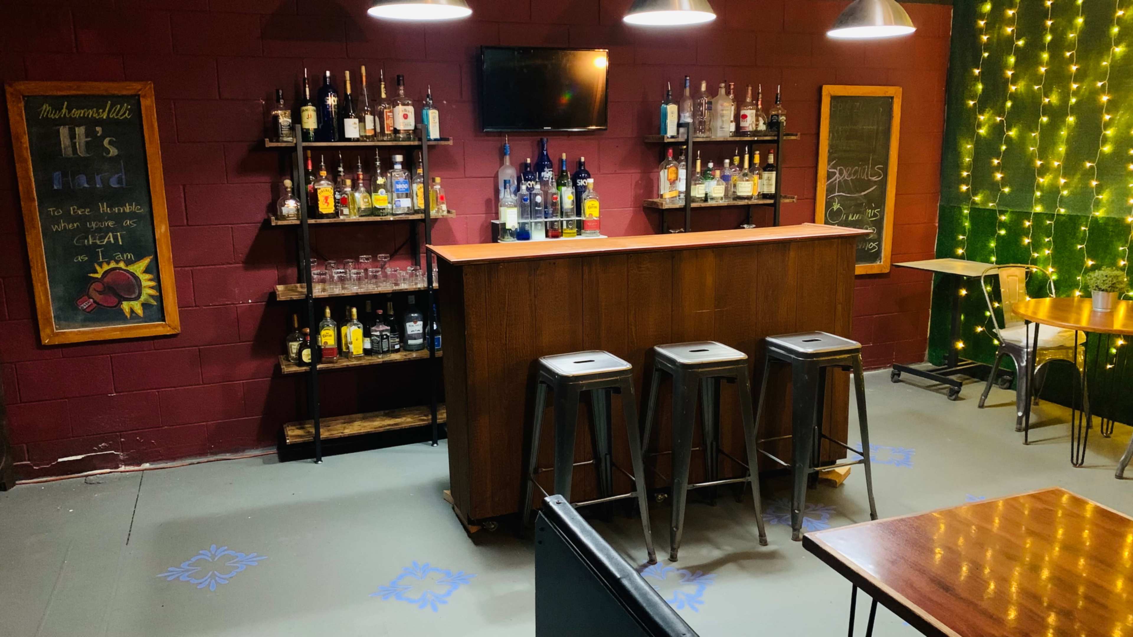 A bar area with a wooden counter and three metal stools, surrounded by shelves holding various bottles and a chalkboard listing specials.