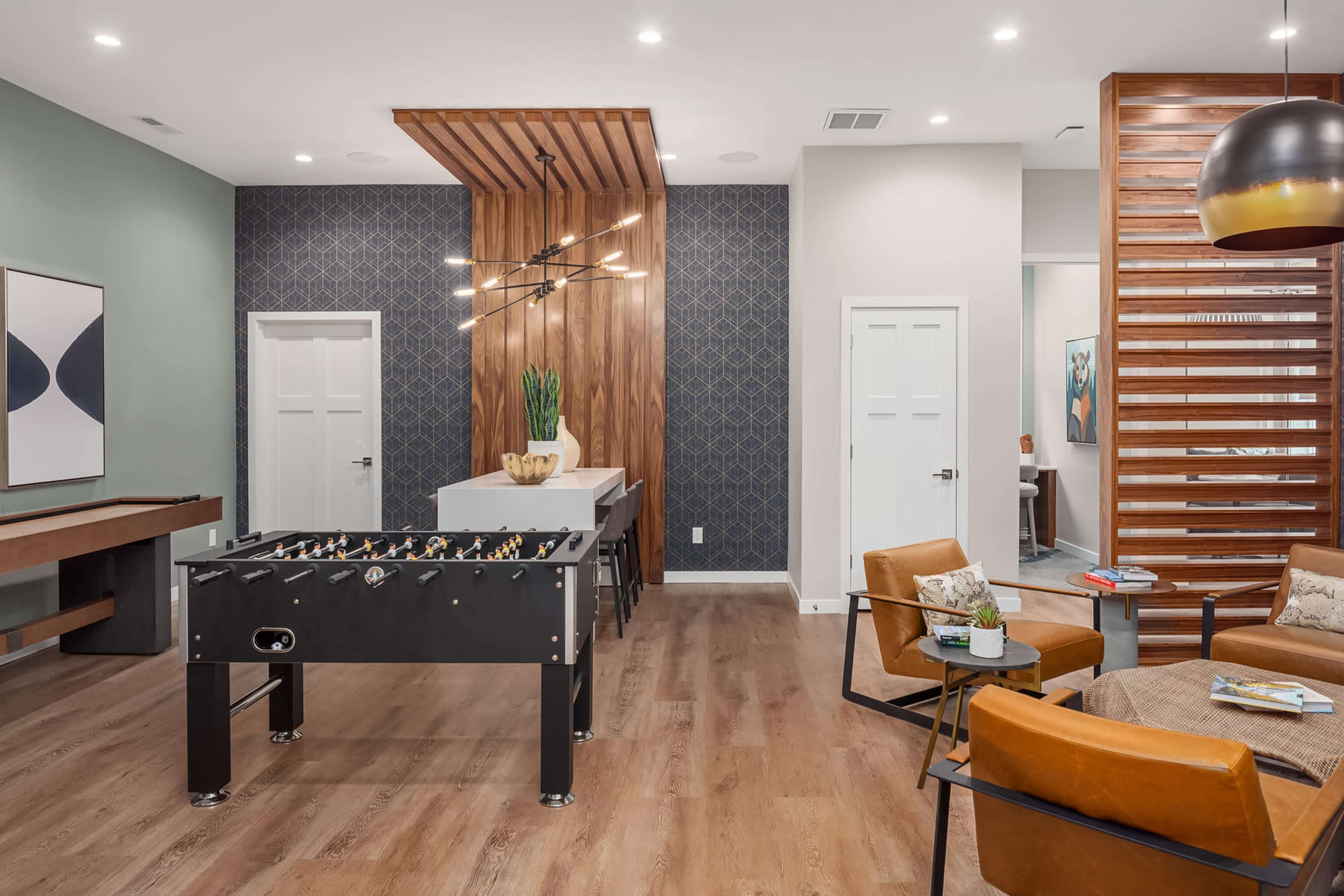 A modern lounge area featuring a foosball table, a dining setup, and cozy seating, with decorative lighting and wall patterns.