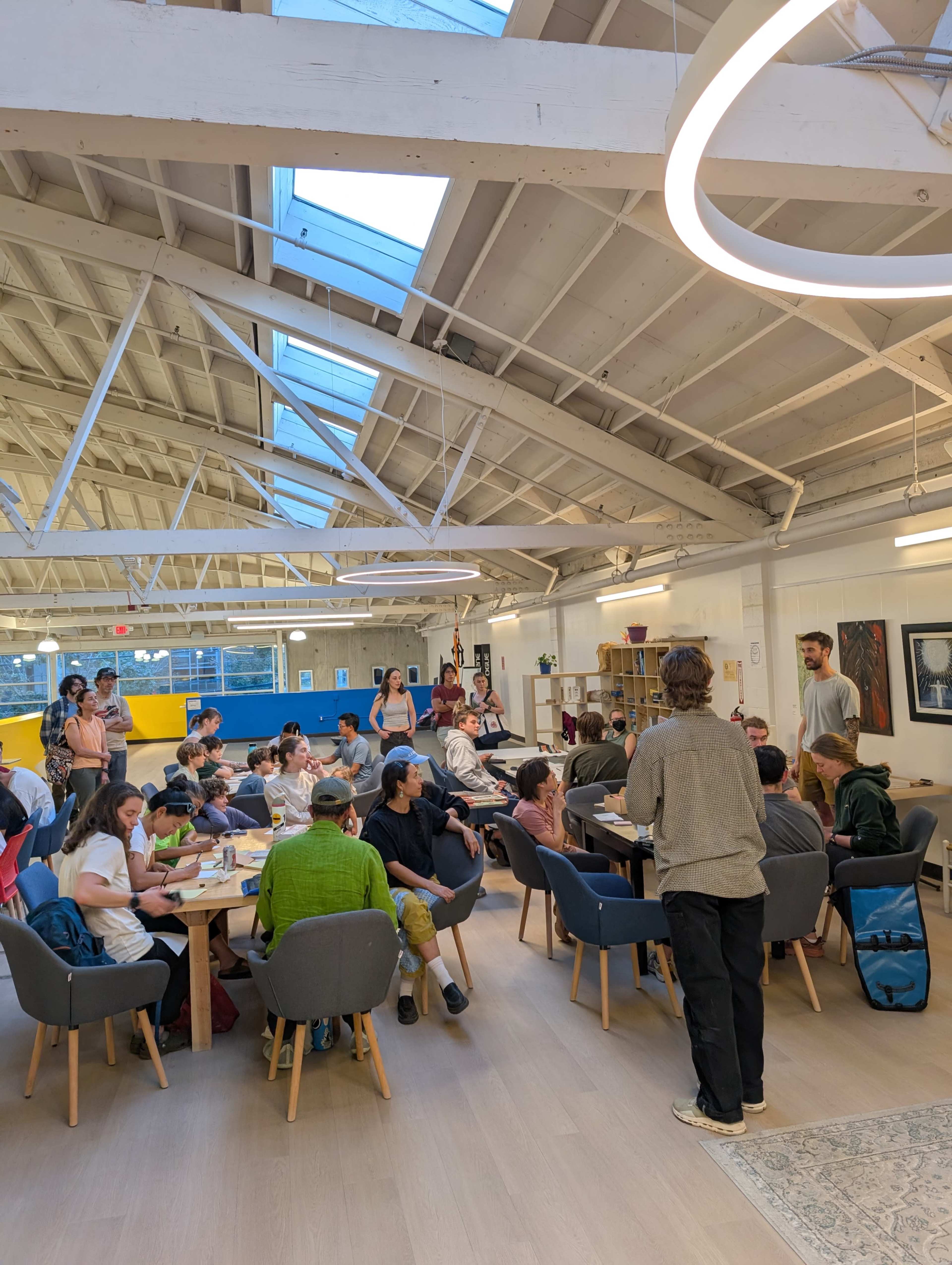 A large group of people are seated around tables in a brightly lit, open space with modern furnishings and high ceilings.