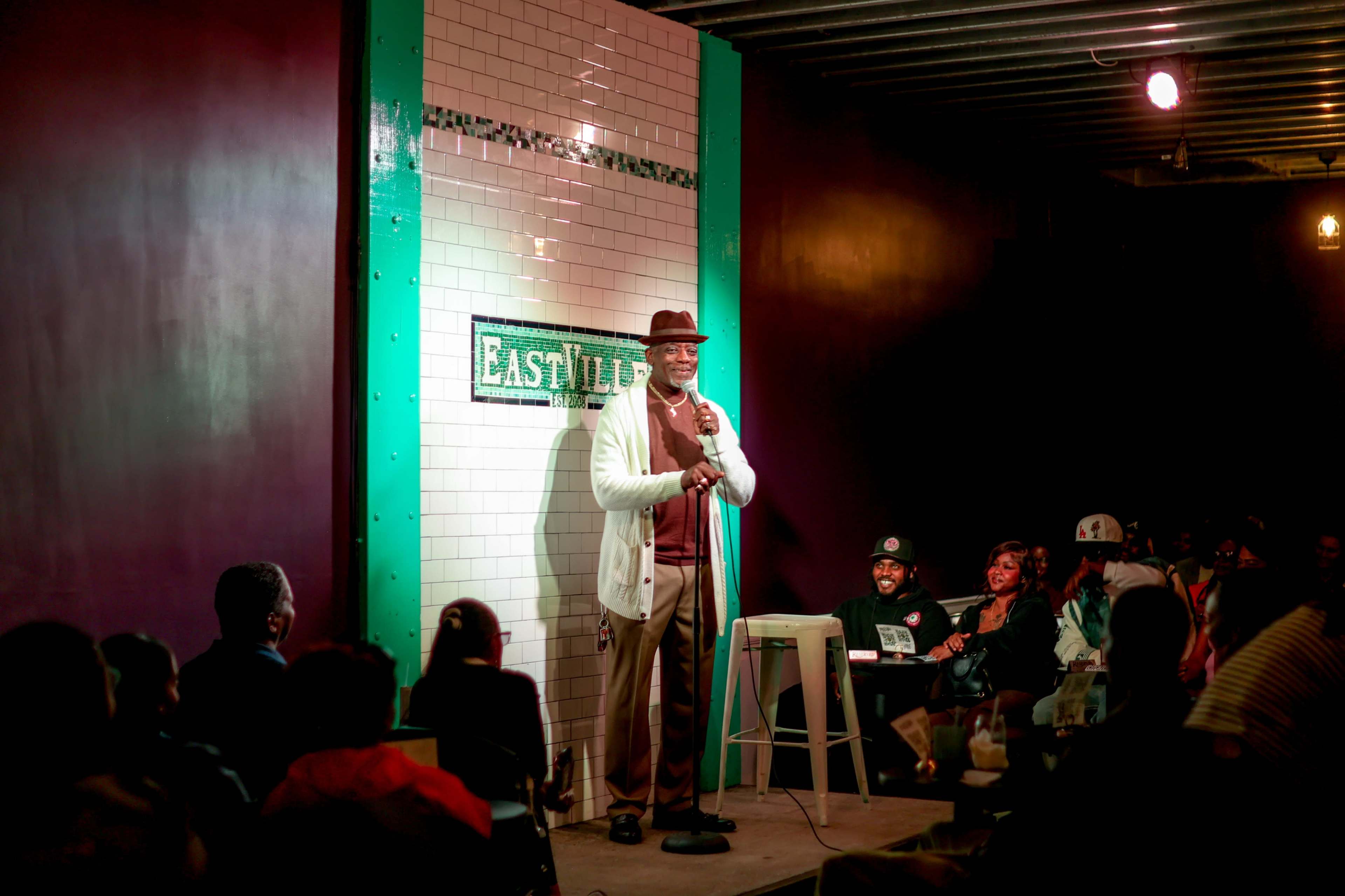 A comedian stands on stage with a microphone, performing in a small, dimly lit comedy club.