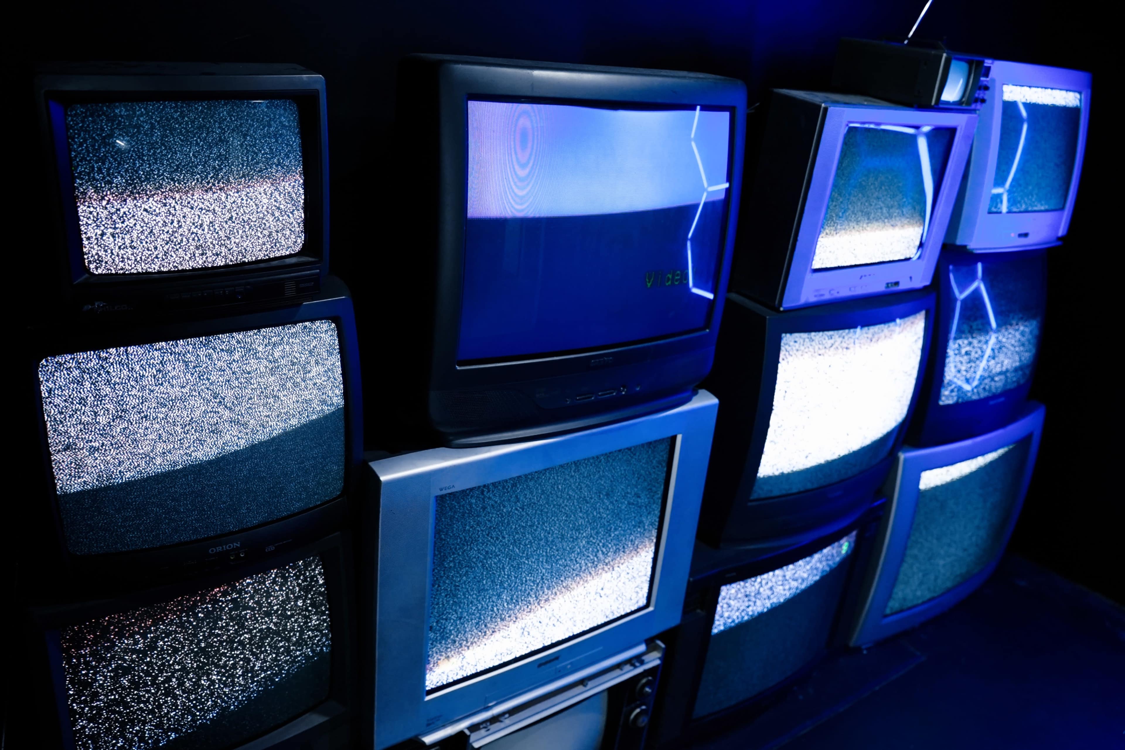 A collection of old televisions displaying static is arranged against a dark wall, illuminated by blue lighting.