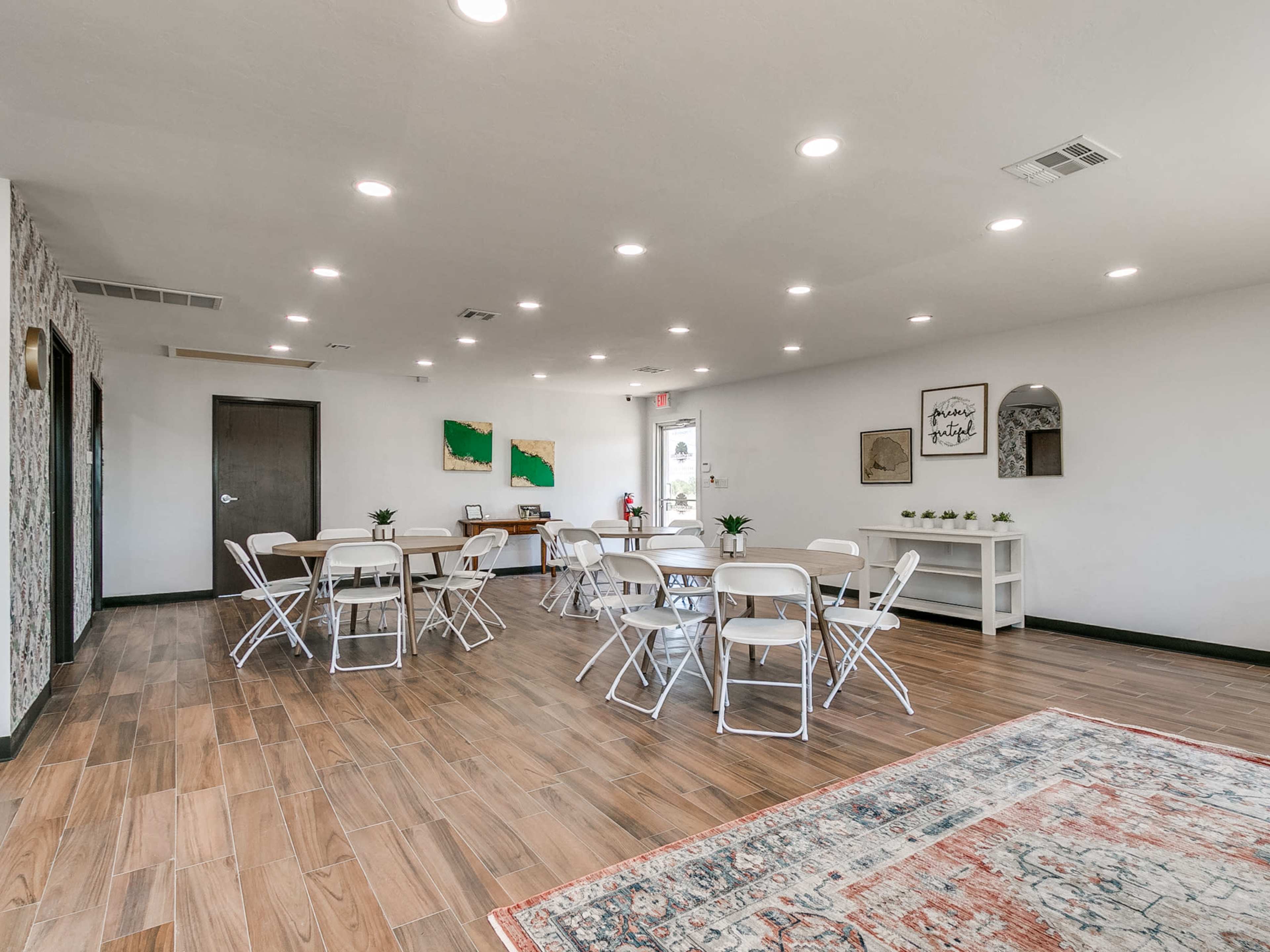 A spacious, well-lit room with several tables and chairs arranged for a gathering, featuring light wood flooring and decorative wall art.