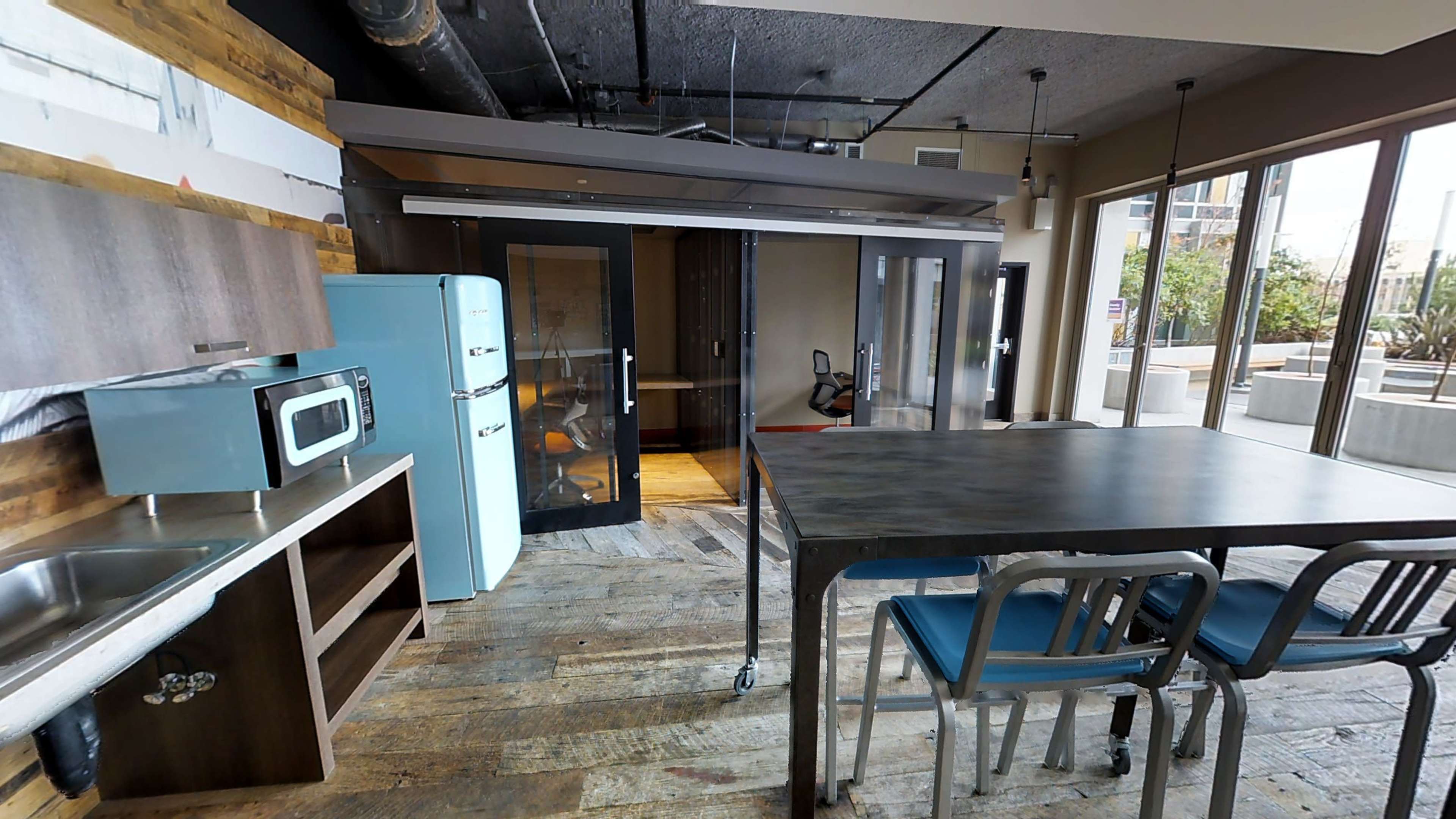 The image shows a modern kitchen area with a blue refrigerator, a microwave, and a large table surrounded by chairs, all set in an open space with large windows.