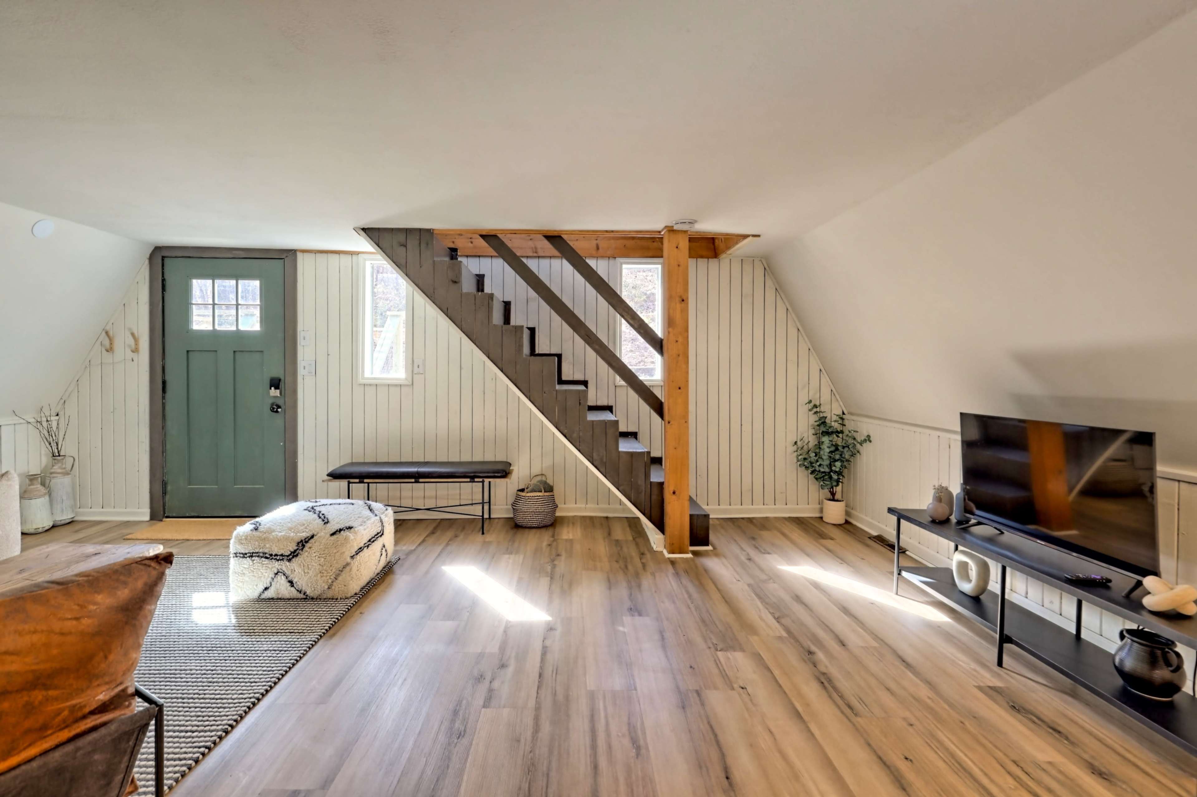 A well-lit living room with a staircase, a green door, and a television set against a wooden floor.