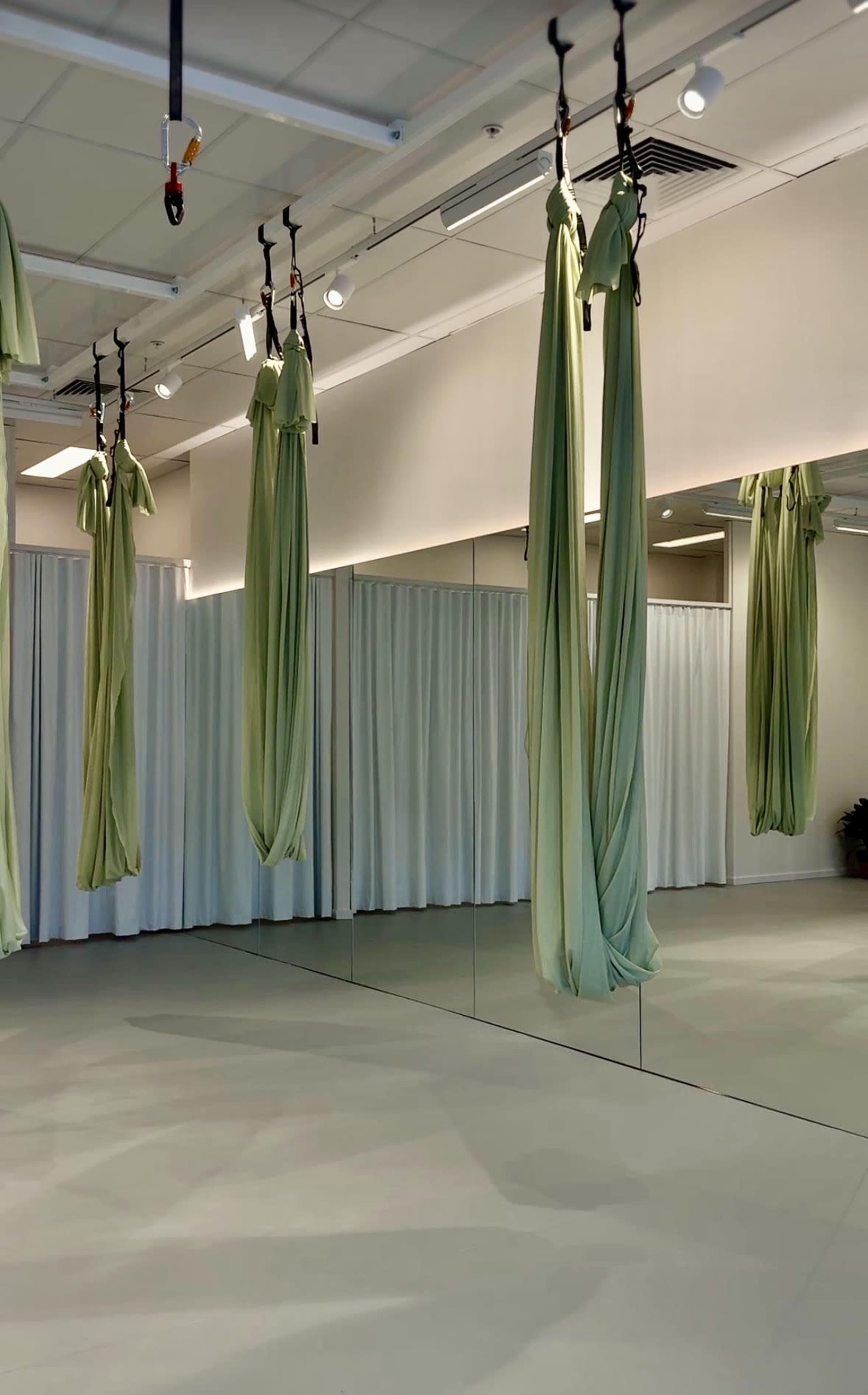 The image shows an aerial yoga studio featuring green silk hammocks suspended from the ceiling, with large mirrors and white curtains lining the walls.