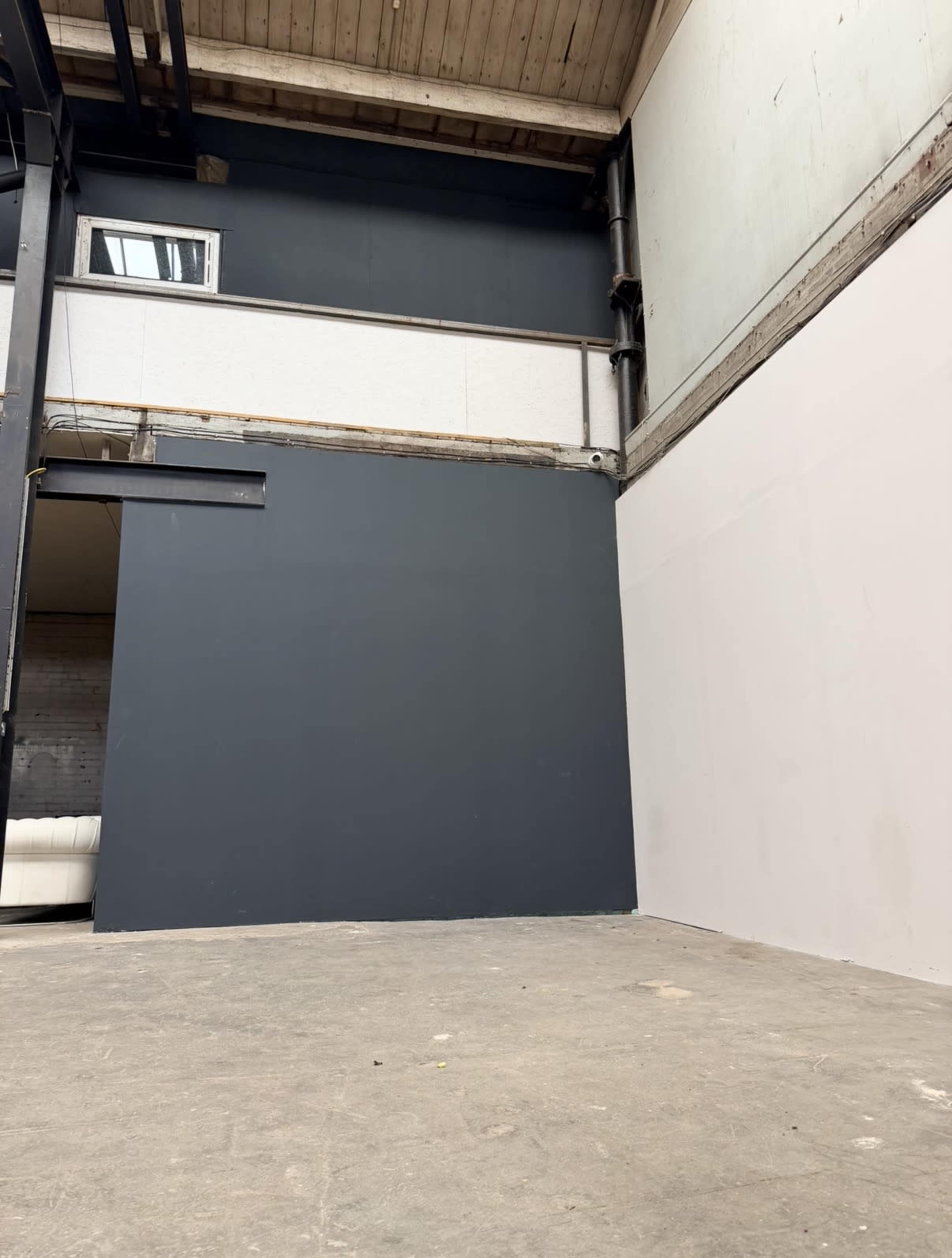 The image shows an industrial interior space with a gray wall, exposed beams, and an unfinished concrete floor.