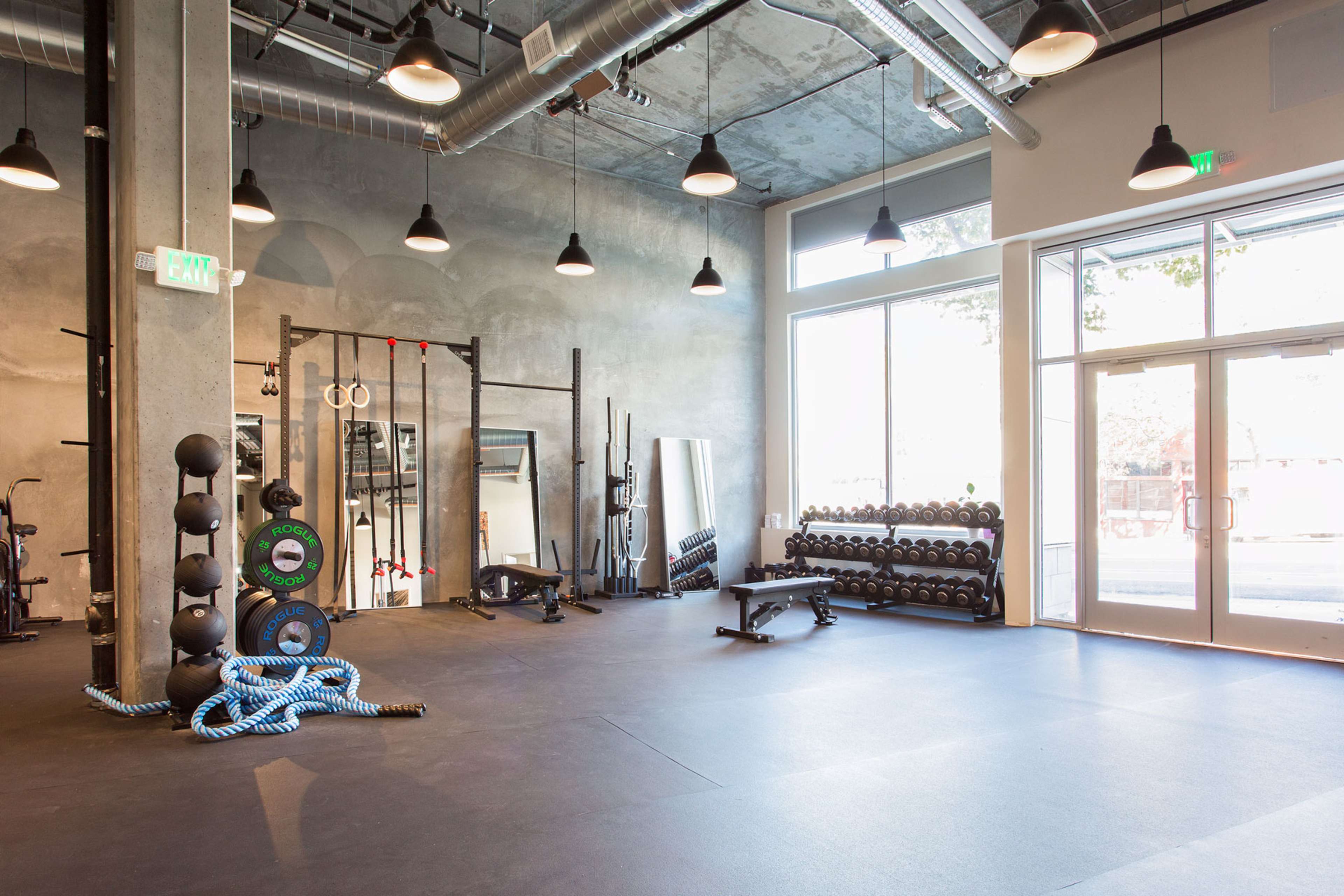 A spacious gym interior featuring various workout equipment, including dumbbells, benches, and resistance bands, with large windows allowing natural light to enter.