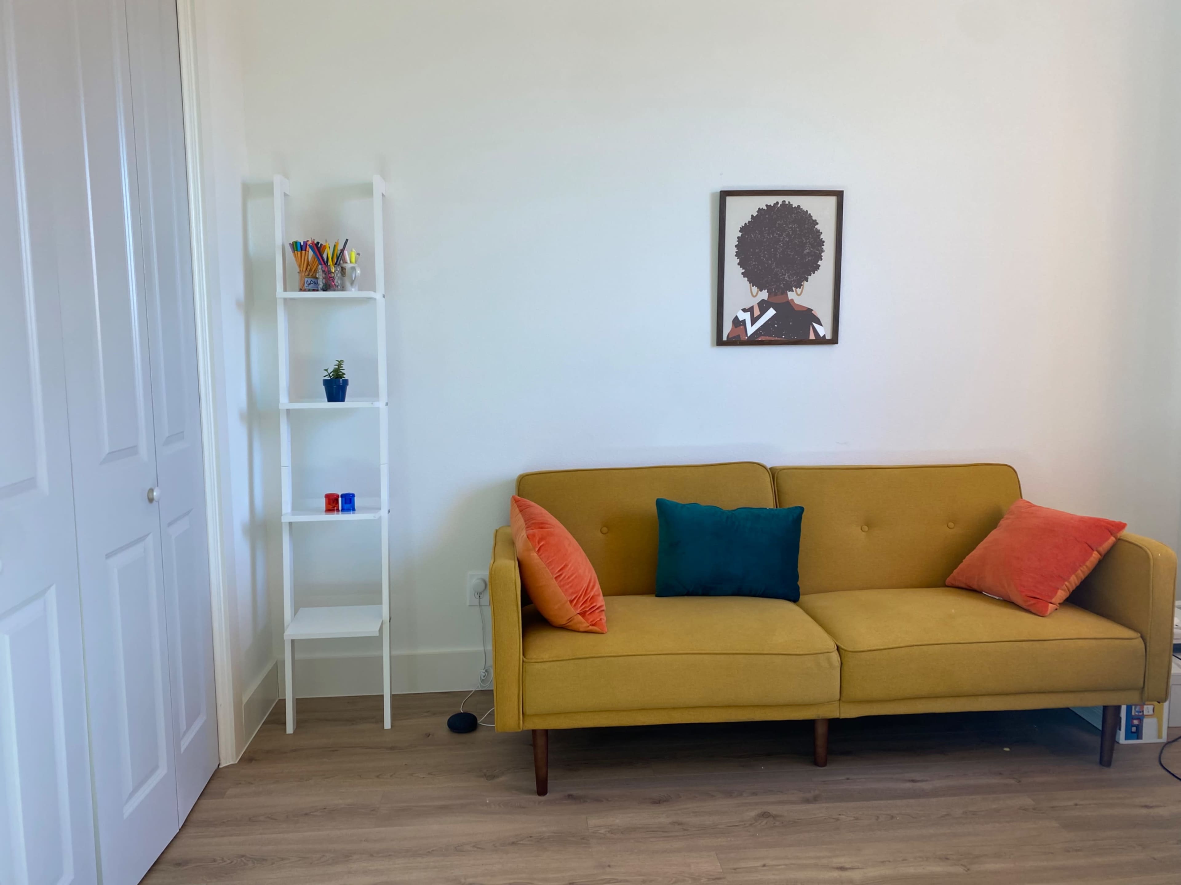 A yellow couch with orange and blue pillows sits against a white wall, next to a white shelving unit with various items and a framed artwork above it.