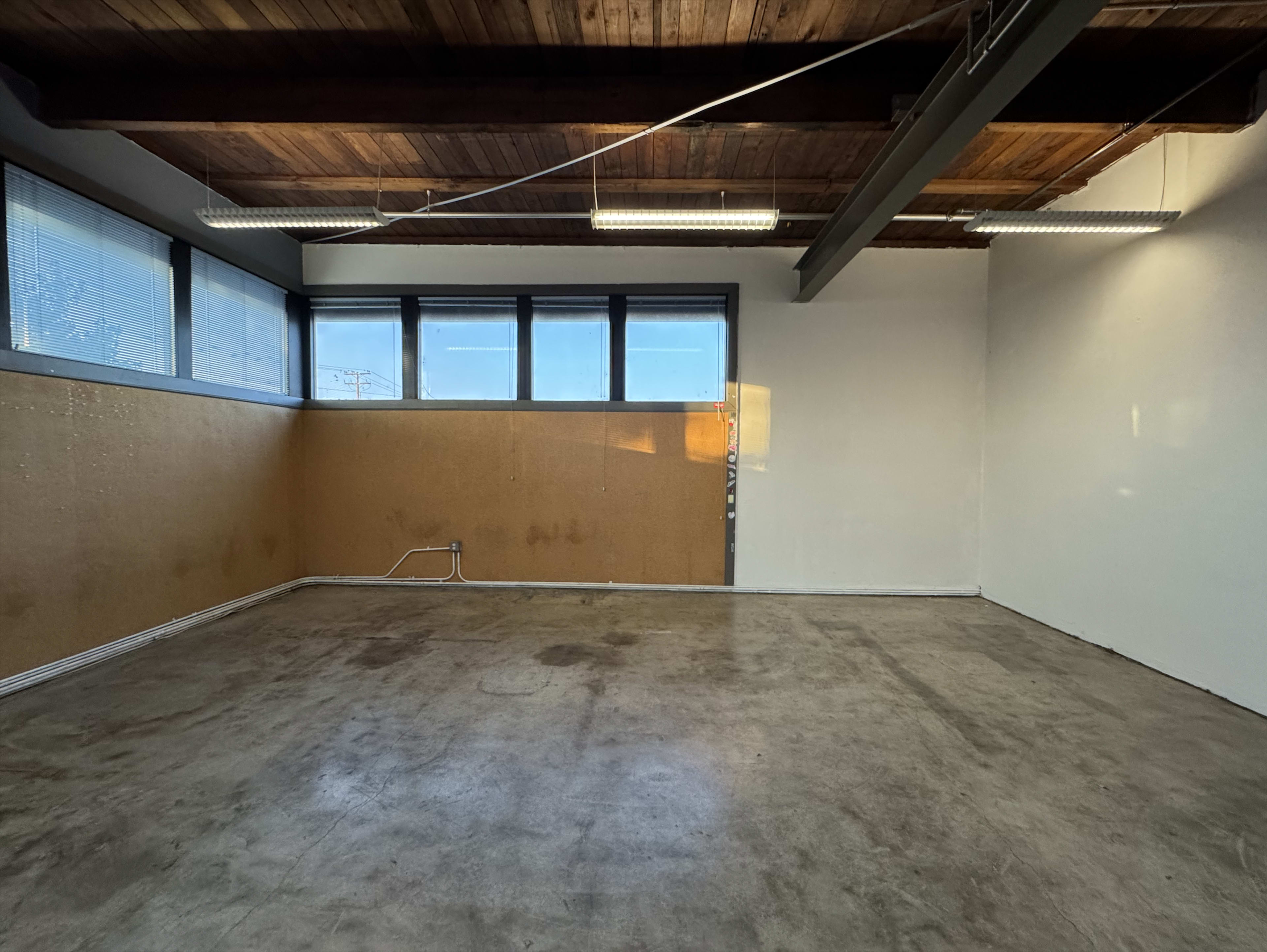 The image shows a spacious, empty room with concrete flooring, large windows, and a wooden ceiling.
