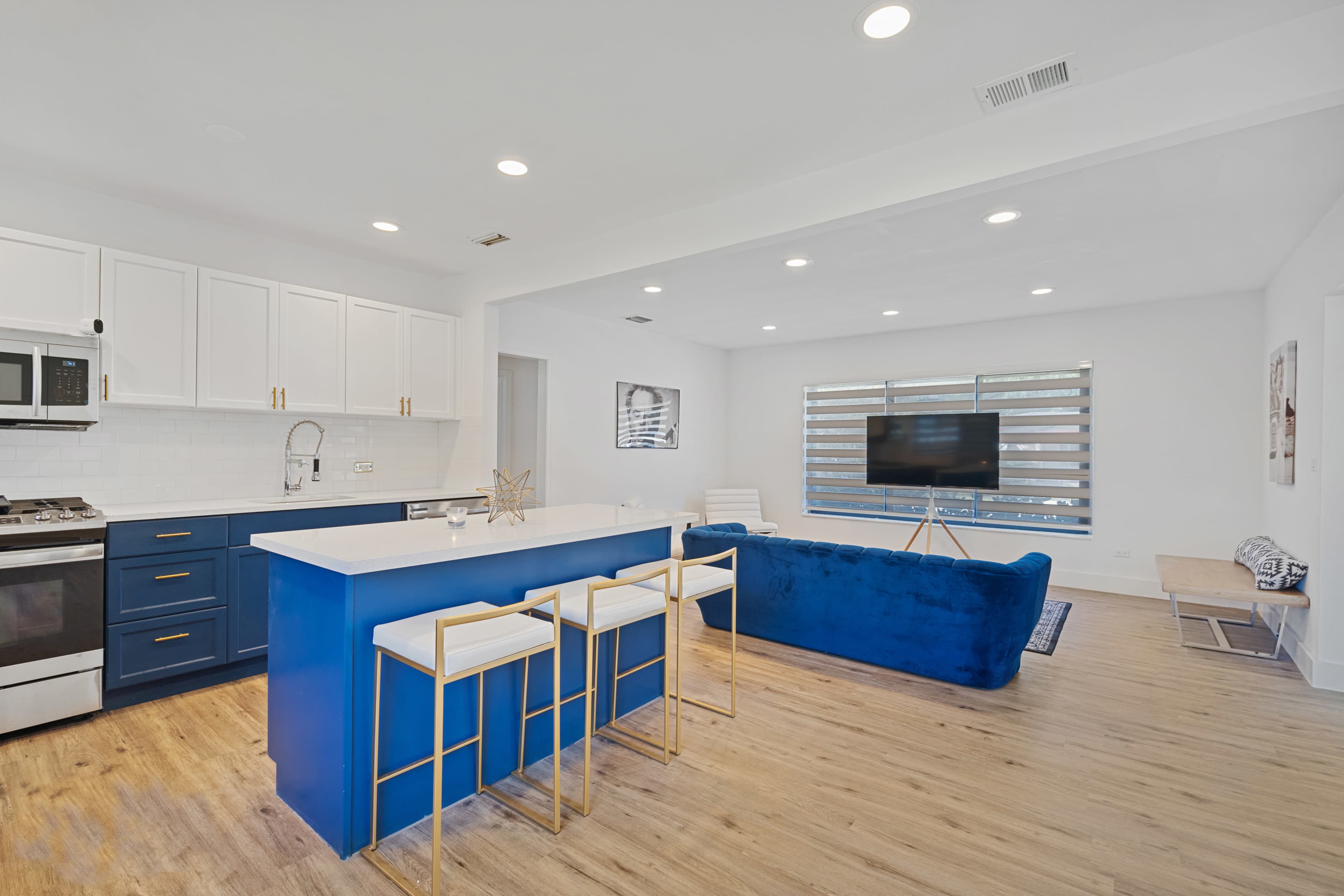 The image shows a modern kitchen and living area featuring a blue island with stools, white cabinetry, and a television on the wall across from a blue couch.