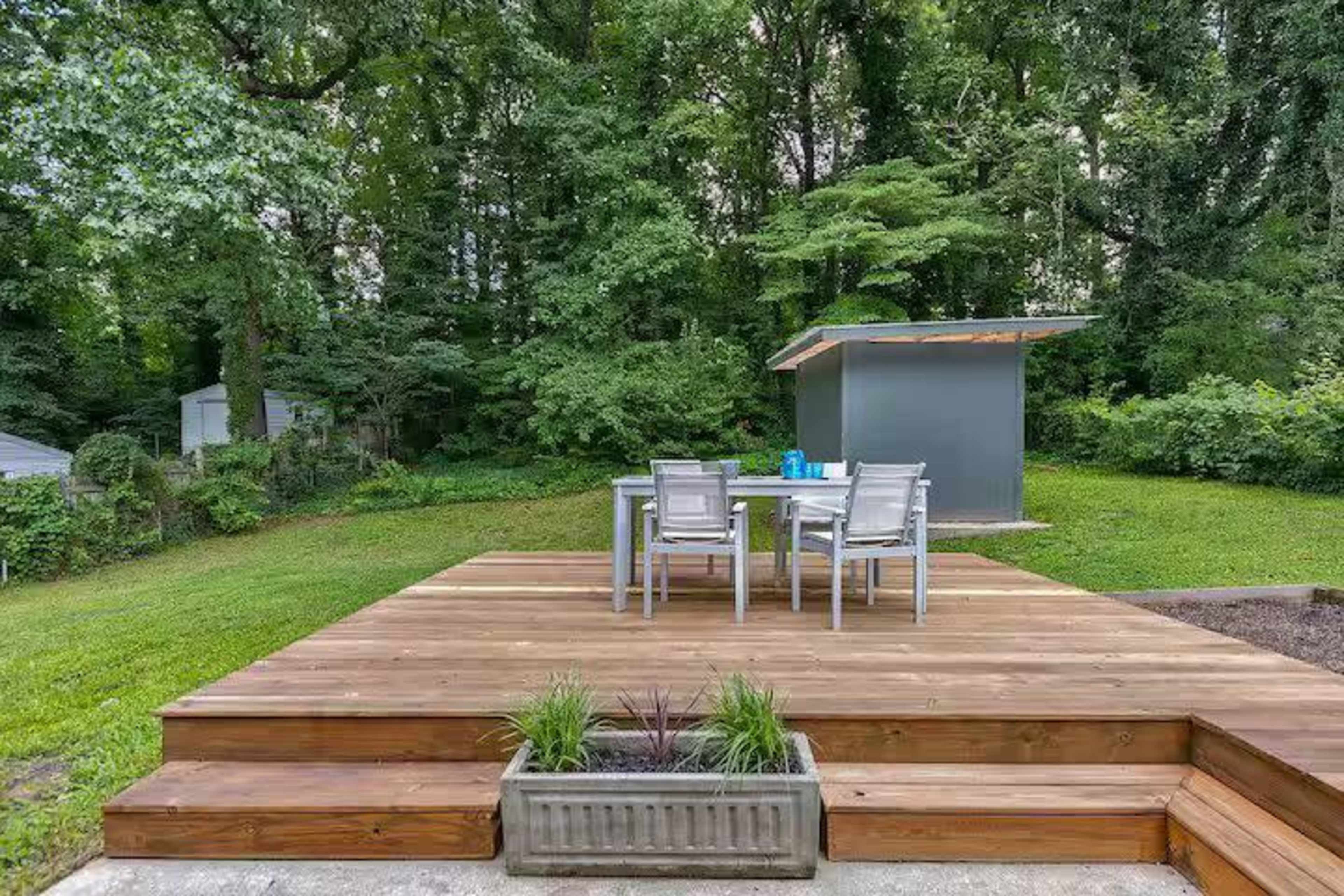 A wooden deck features a table and chairs set against a backdrop of dense greenery and a storage shed.