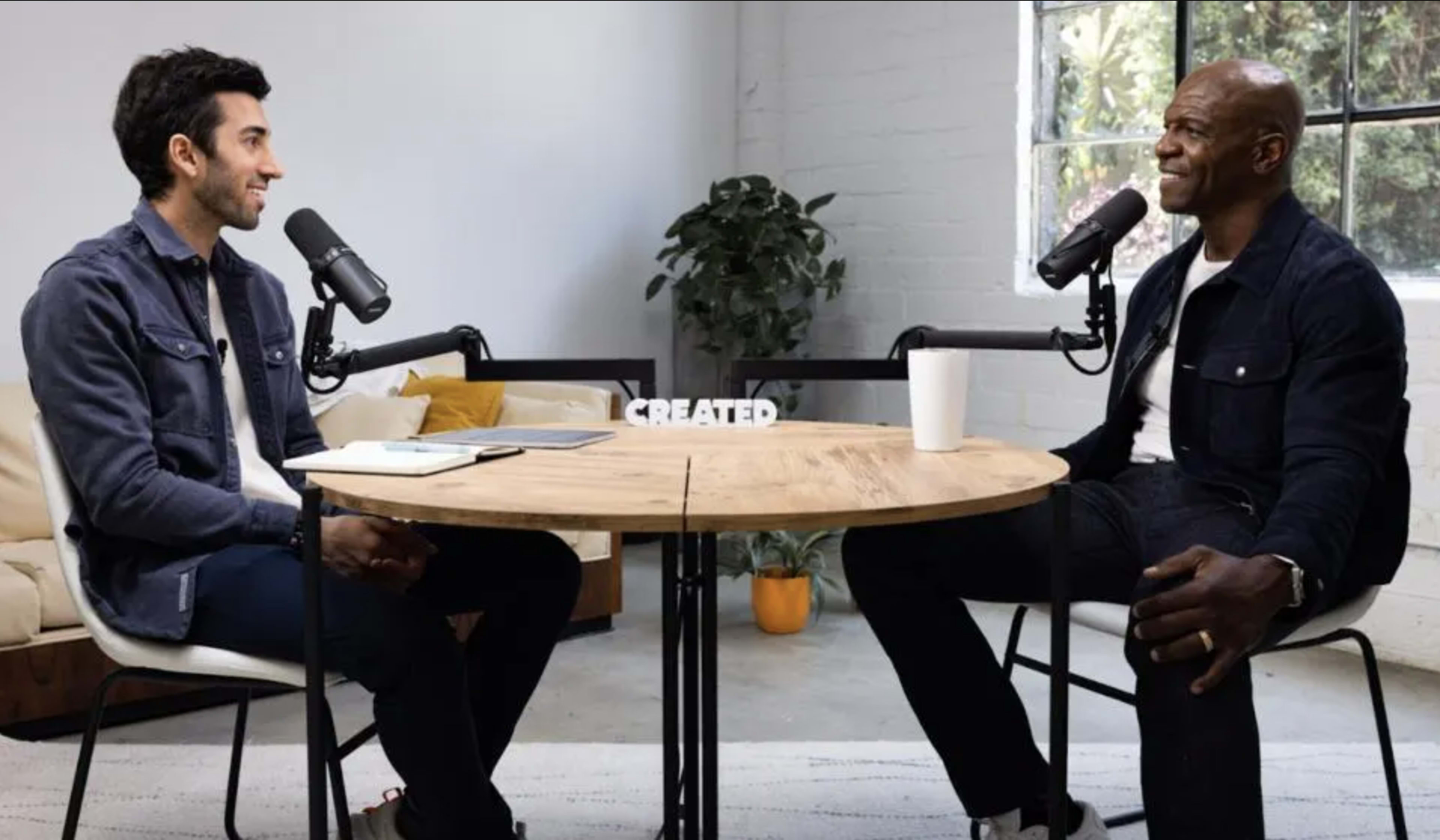 Two men sit at a round table with microphones, engaged in conversation in a bright room featuring a plant and a couch.