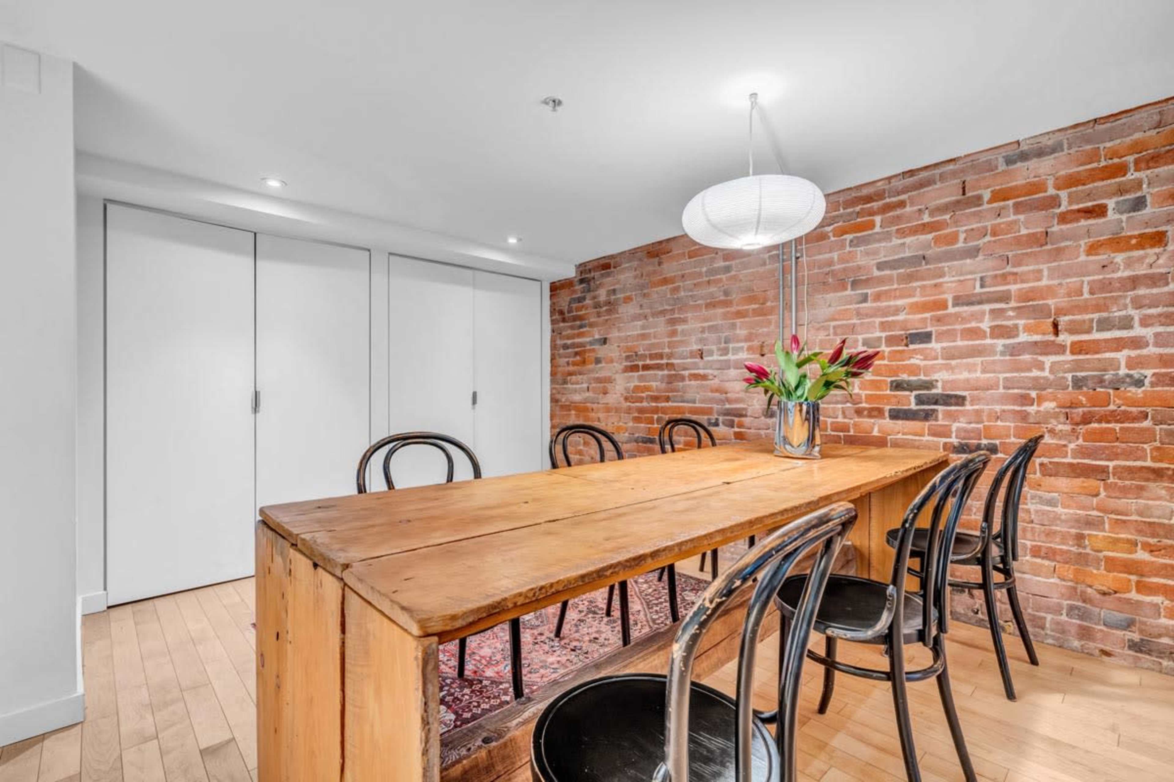 A dining area features a large wooden table with black chairs, set against a brick wall, and is illuminated by a pendant light.