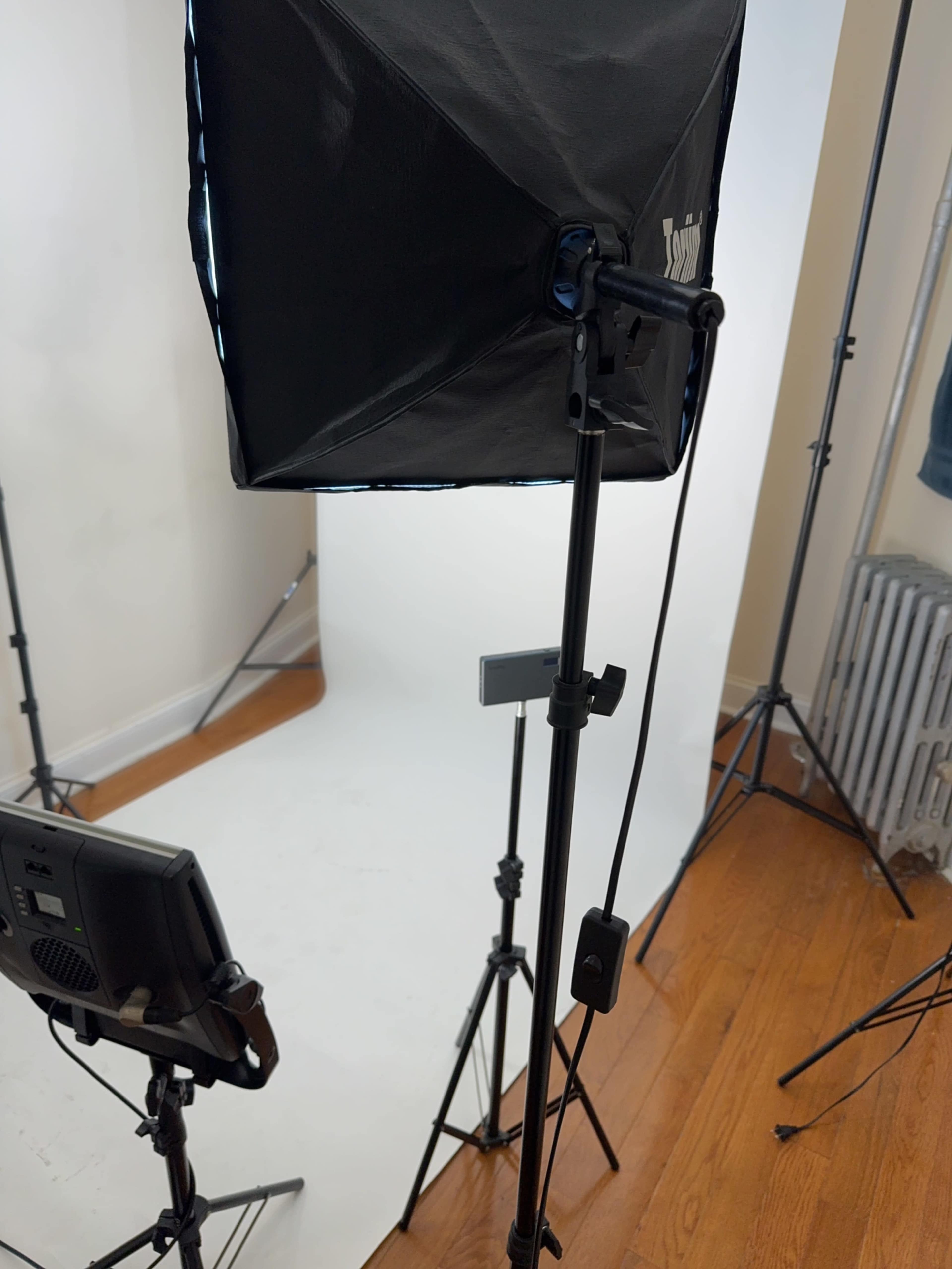 The image shows a photography setup featuring a softbox light, a camera on a tripod, and a plain white backdrop in a room with wooden flooring.