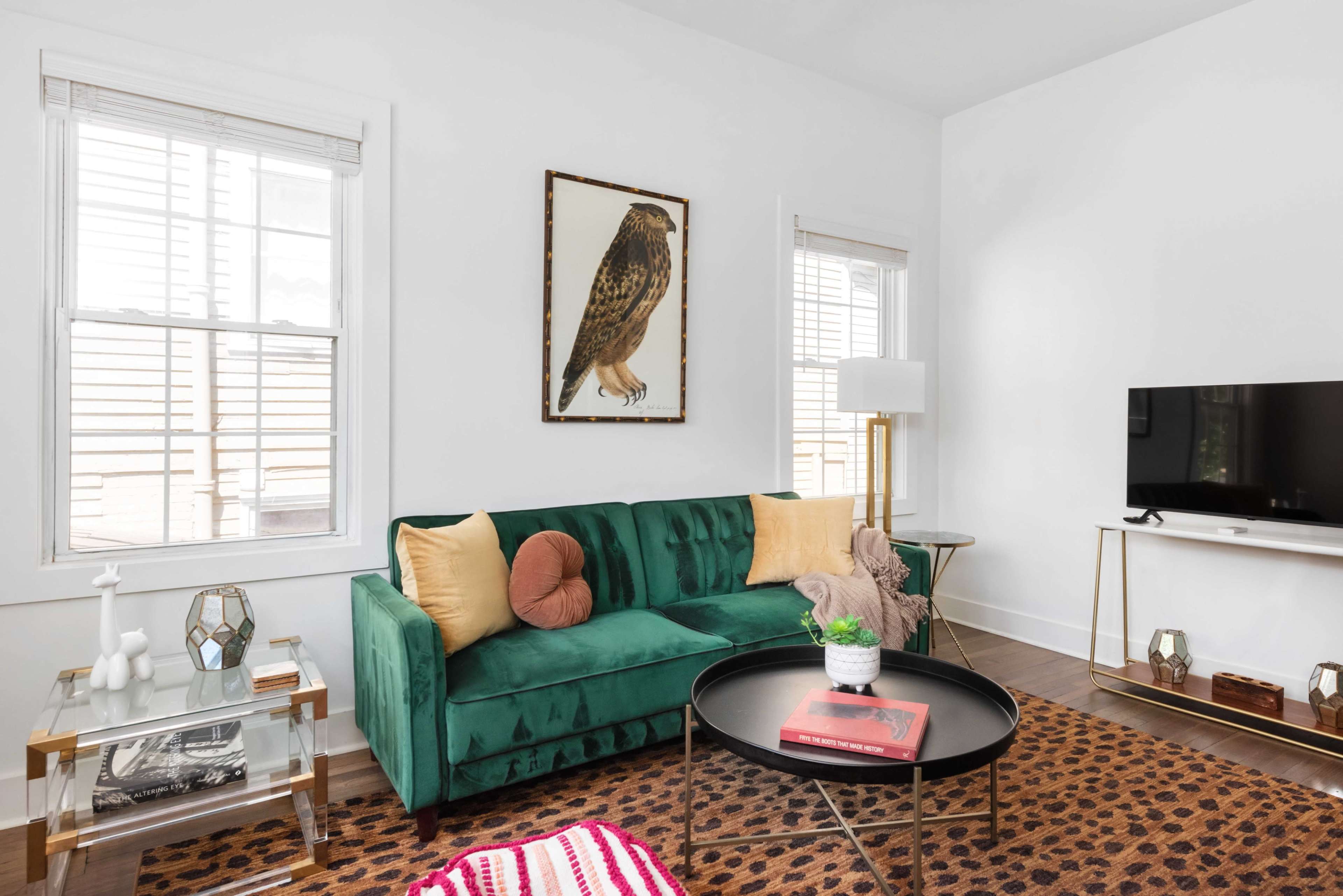 The image shows a bright living room featuring a green velvet sofa, a round coffee table, and a large wall-mounted TV, with light streaming through two windows adorned with blinds.