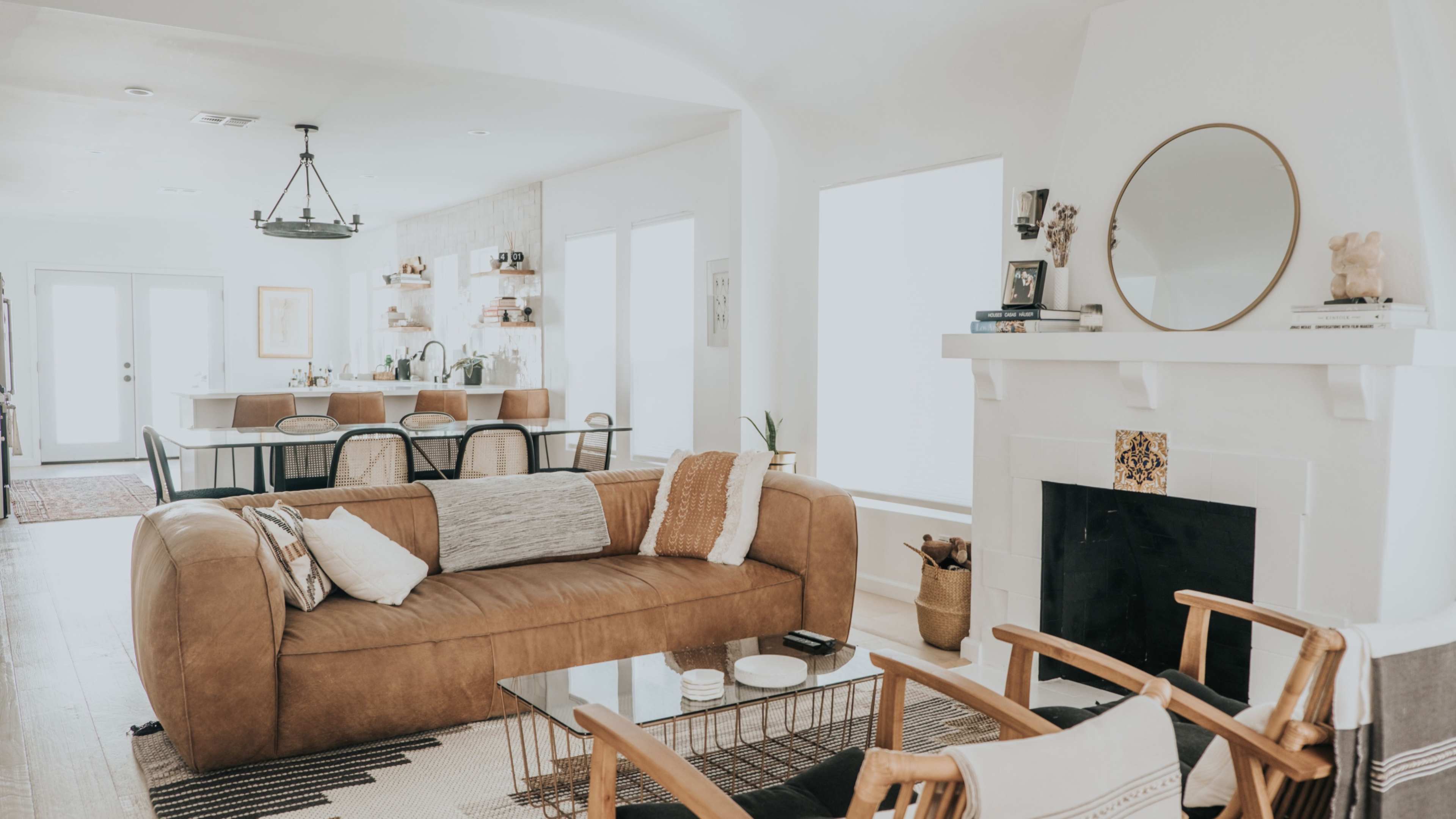 The image shows a modern living room featuring a brown leather sofa, a glass coffee table, and a fireplace with decorative elements, leading to a dining area in the background.